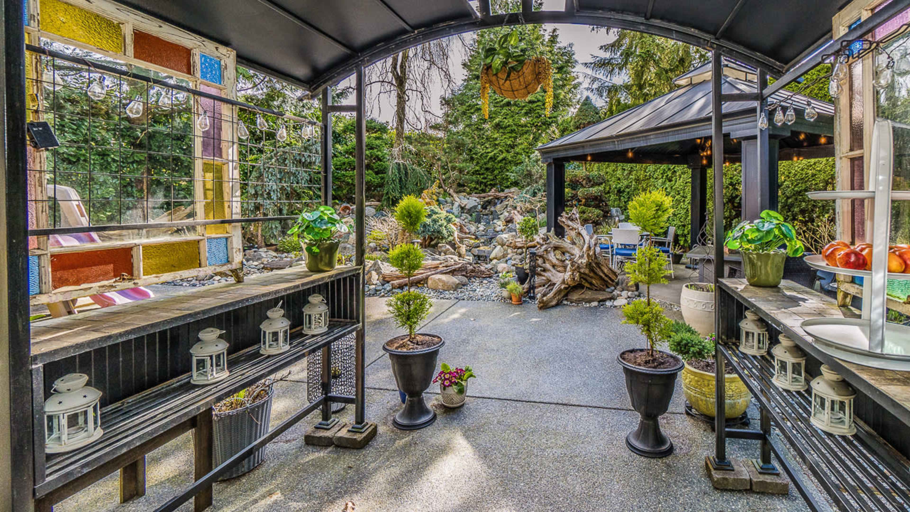 The image shows a landscaped outdoor area featuring a gazebo, decorative plants in pots, and a stone pathway surrounded by various greenery.