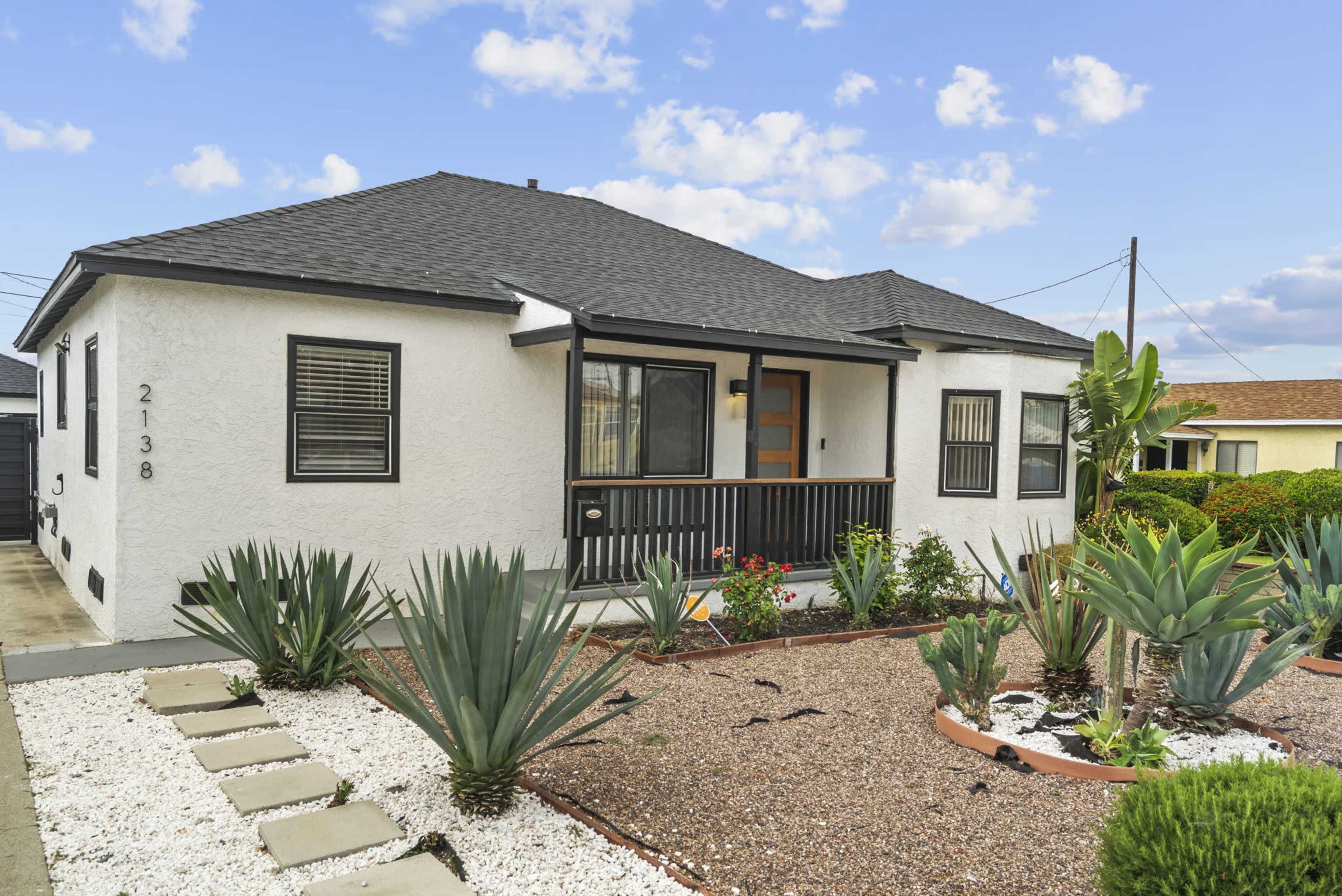 The image shows a modern single-story house with a white exterior, a black roof, and a landscaped front yard featuring various plants and decorative stones.