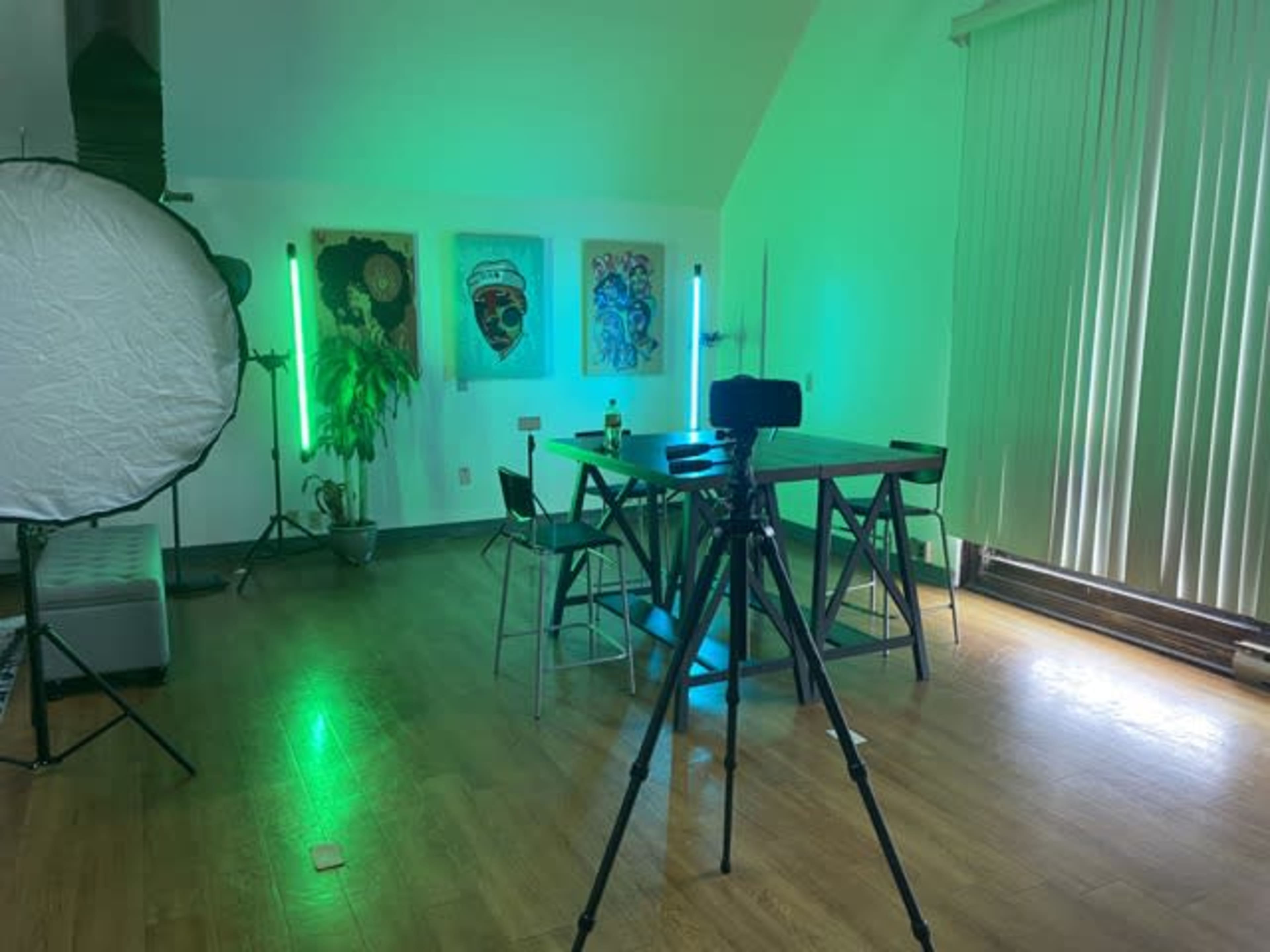 A room is equipped with a tripod and green studio lights, featuring a table surrounded by chairs, and art on the walls.