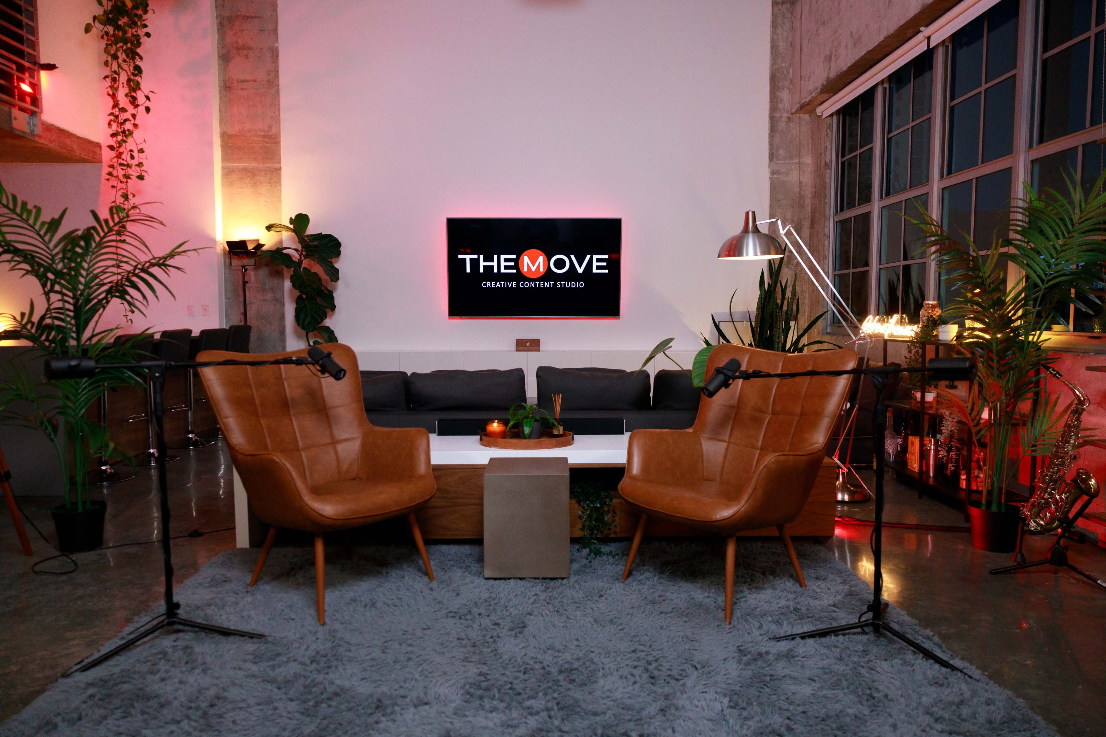 The image shows a modern creative content studio featuring two brown leather armchairs facing a white coffee table, with a large screen displaying "THE MOVE" in the background and plants adorning the space.