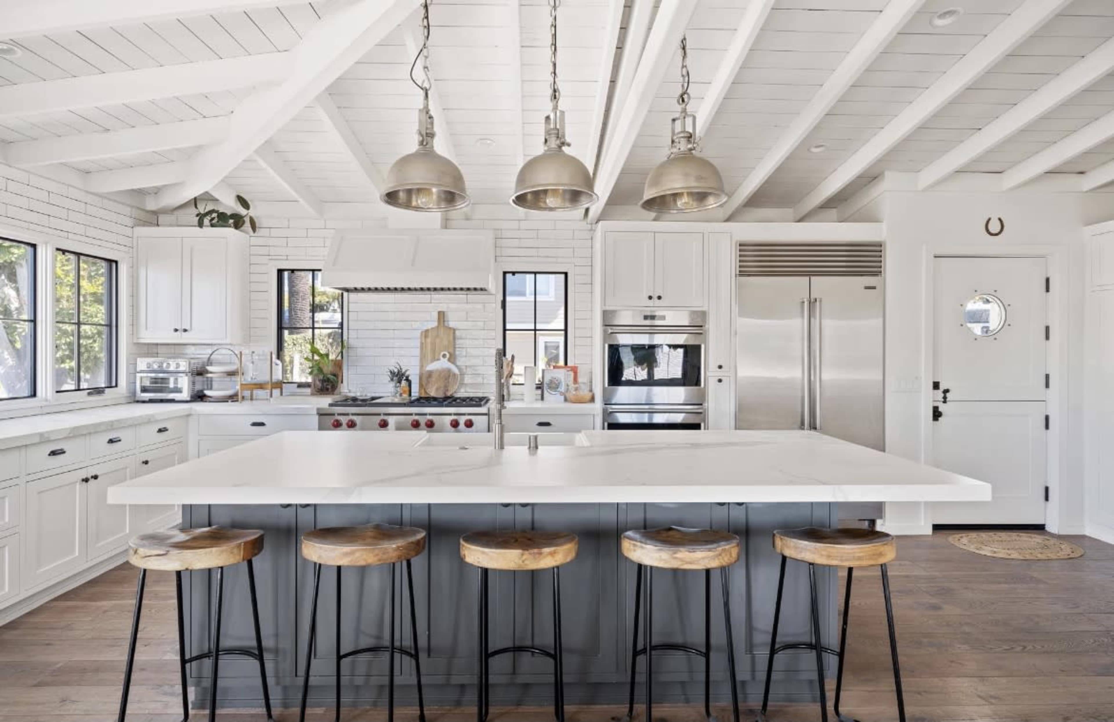 A modern kitchen with a white ceiling, wooden beams, a large island with bar stools, and stainless steel appliances.