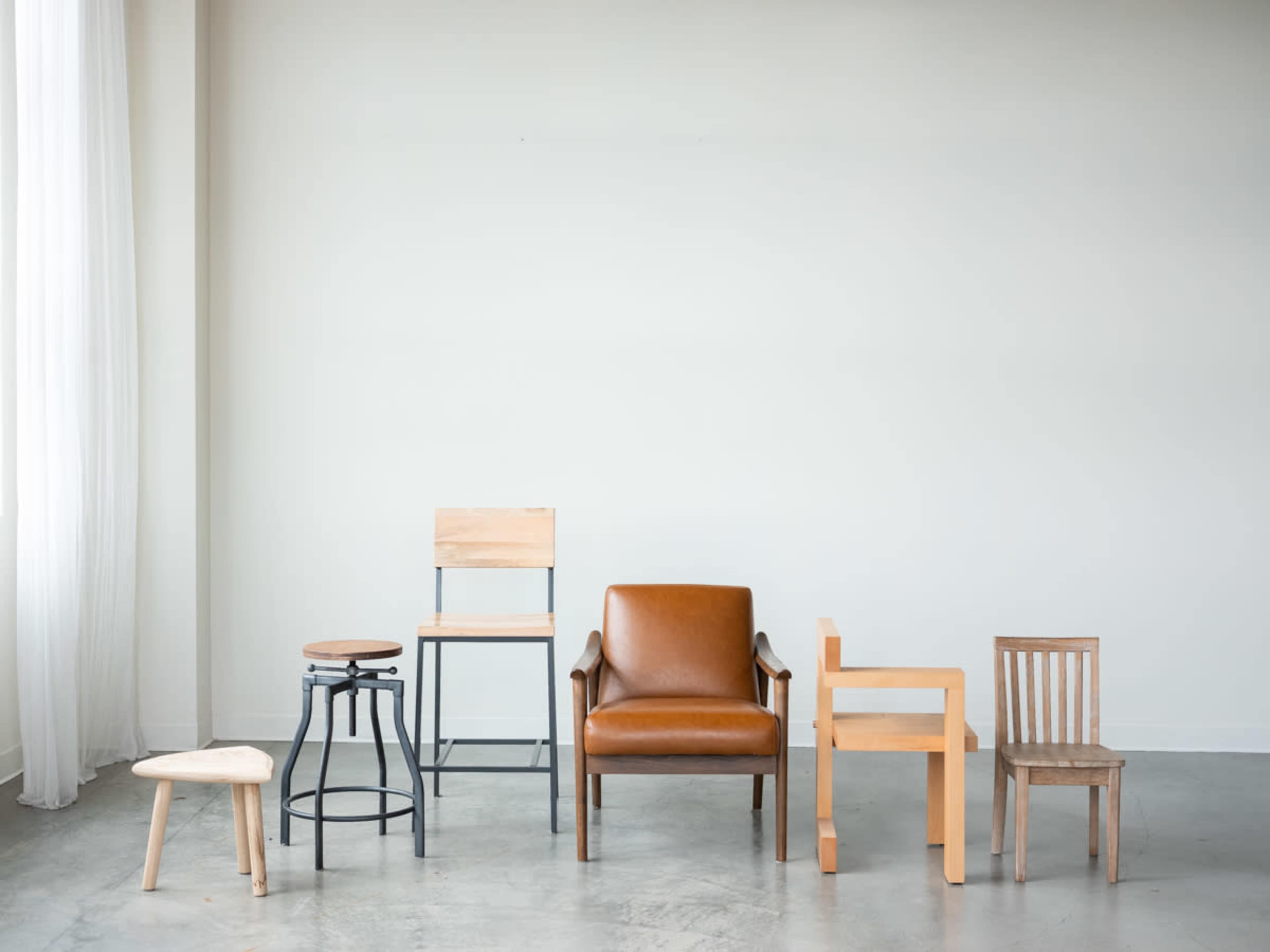 The image shows a collection of various chairs and stools in a minimalist setting against a plain wall.