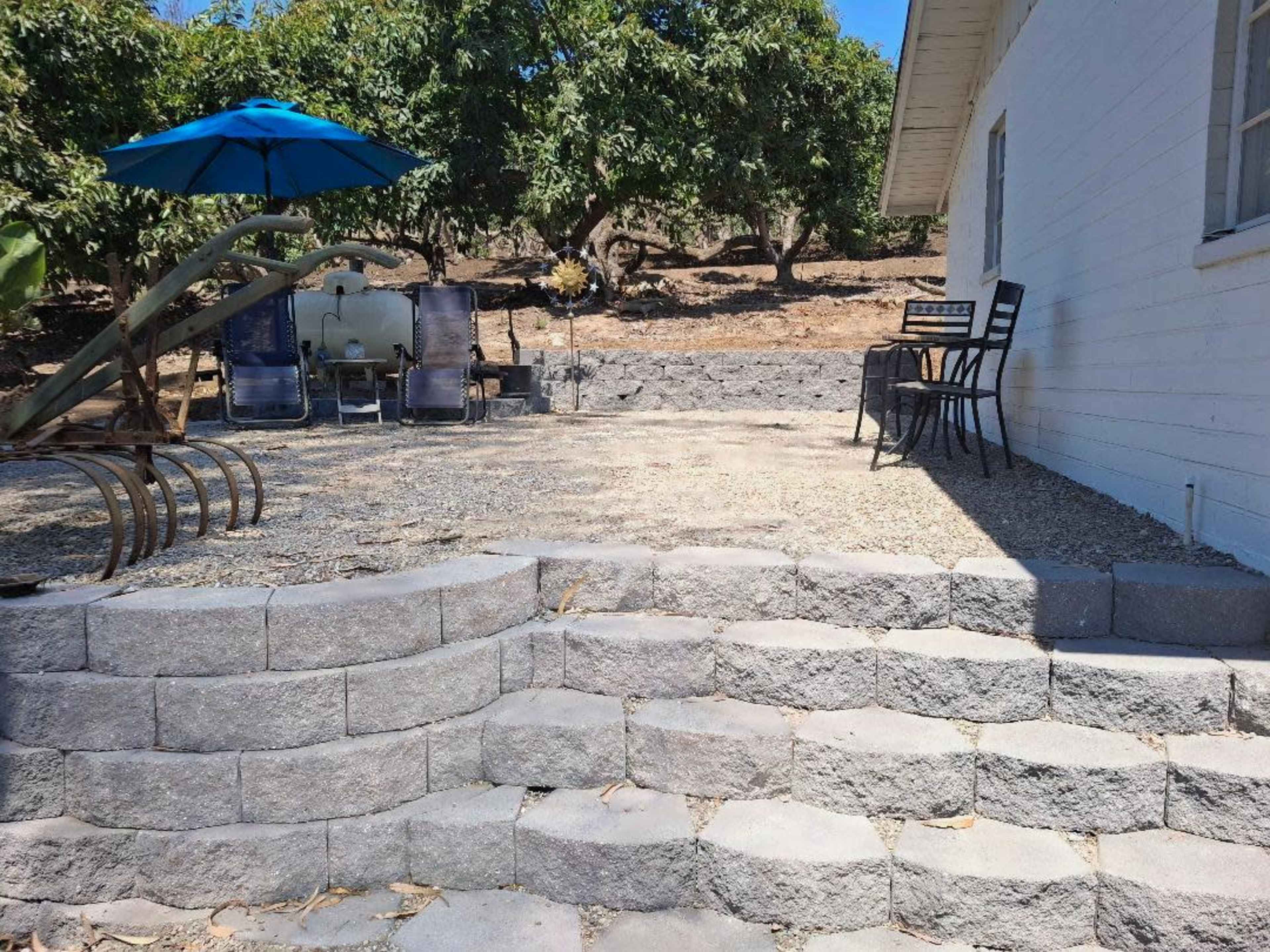 The image shows a landscaped outdoor area featuring stone steps leading up to a patio with chairs, a table, and an umbrella surrounded by trees.