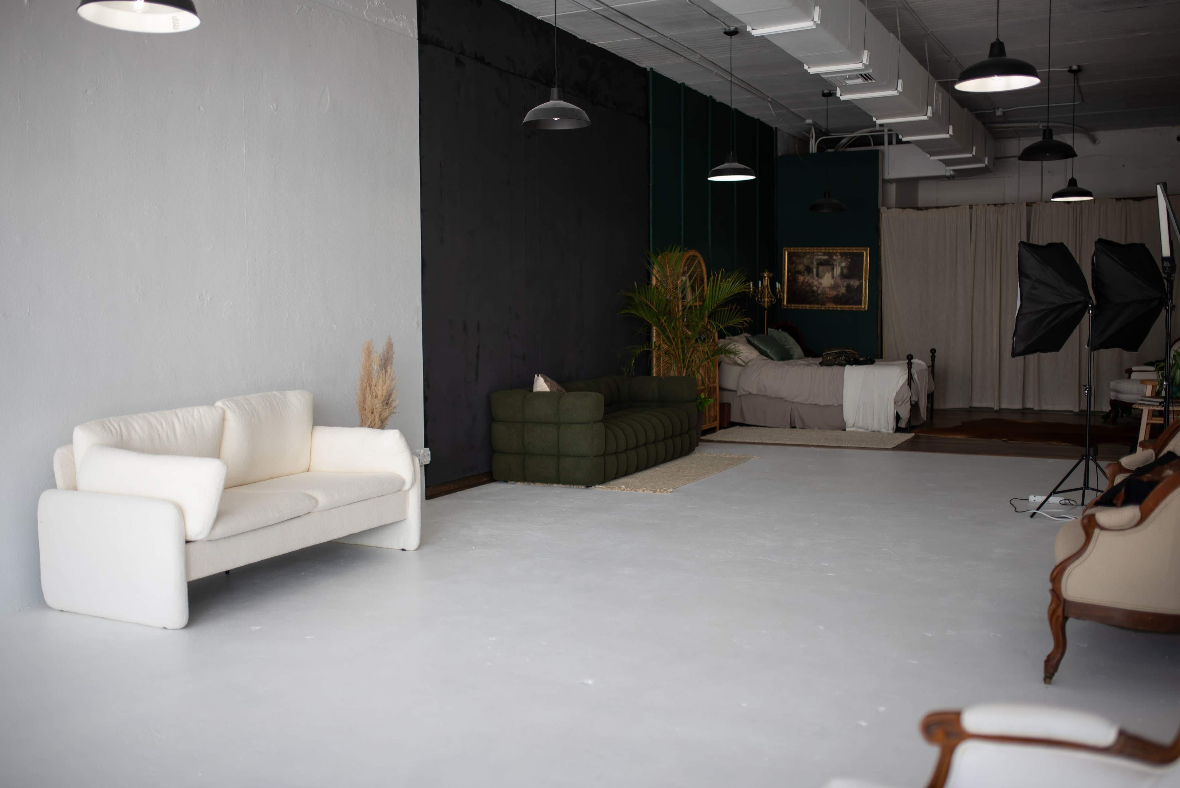 A spacious room divided into two areas, featuring a white sofa and a green couch on one side, and a neatly made bed with decorative plants on the other.