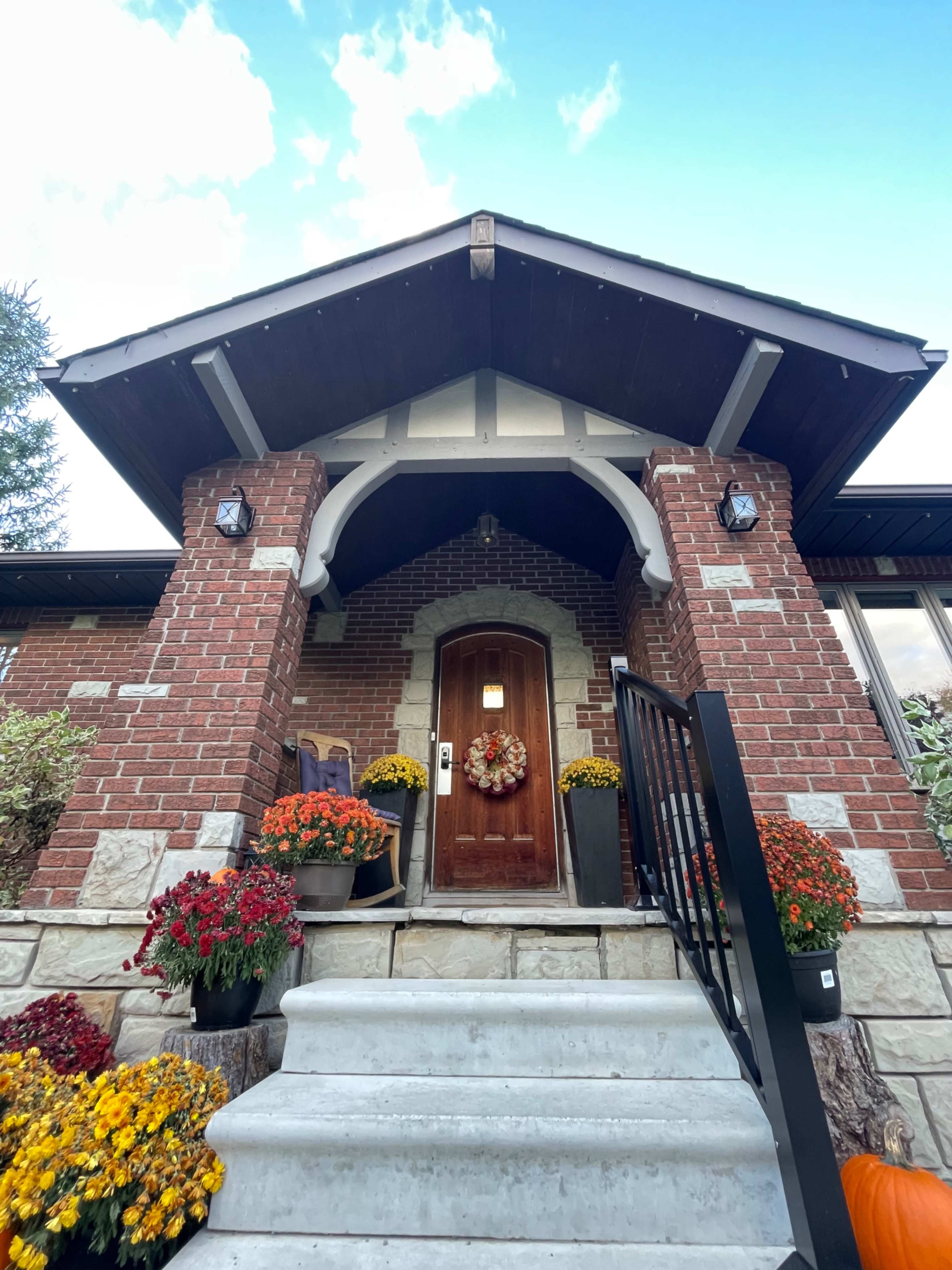 The entrance of a brick house featuring a large wooden door under a decorative archway, flanked by colorful flowerpots and decorative lighting.