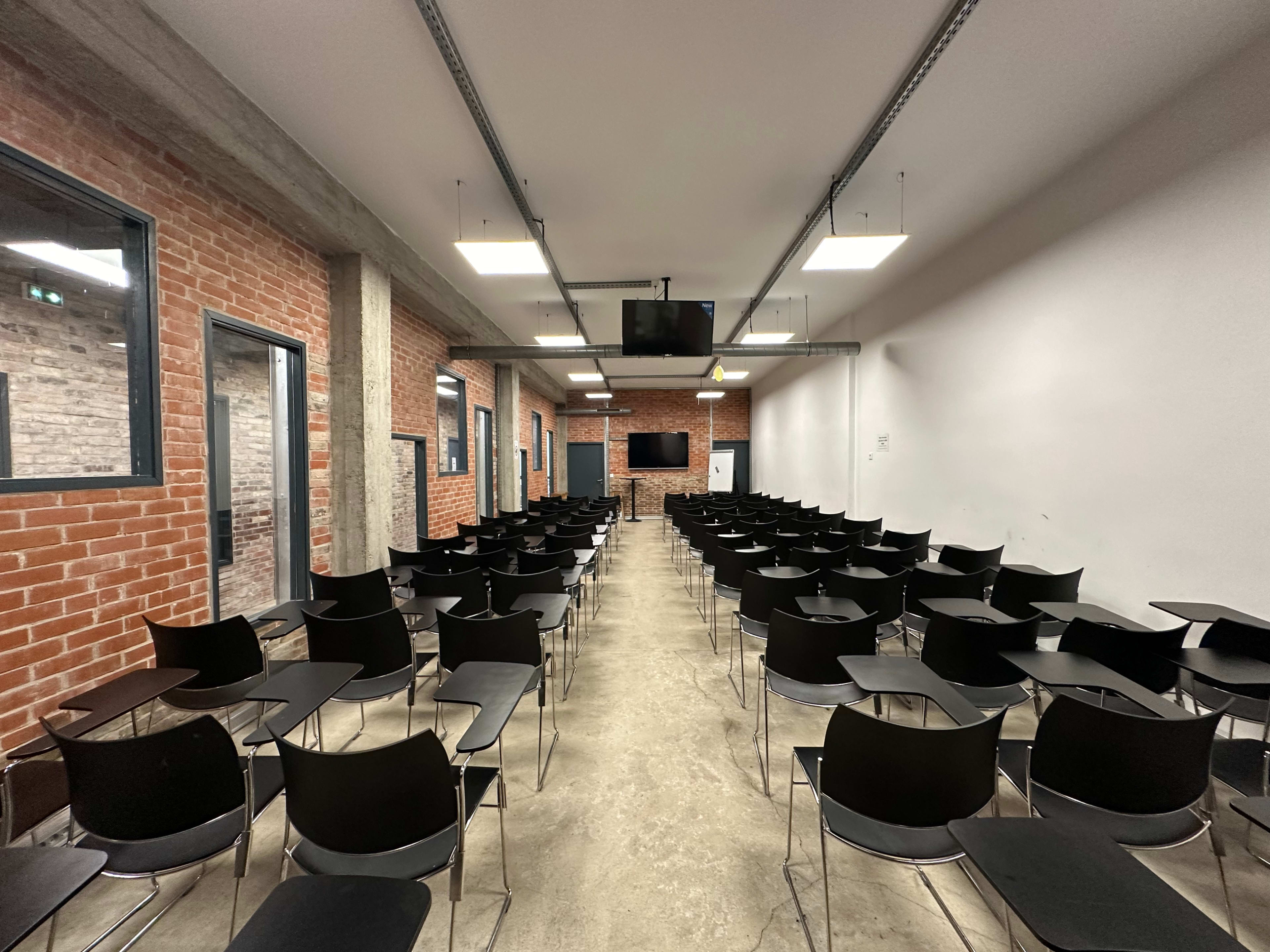 A classroom setup with rows of black chairs arranged facing a screen in a room with exposed brick walls and concrete flooring.