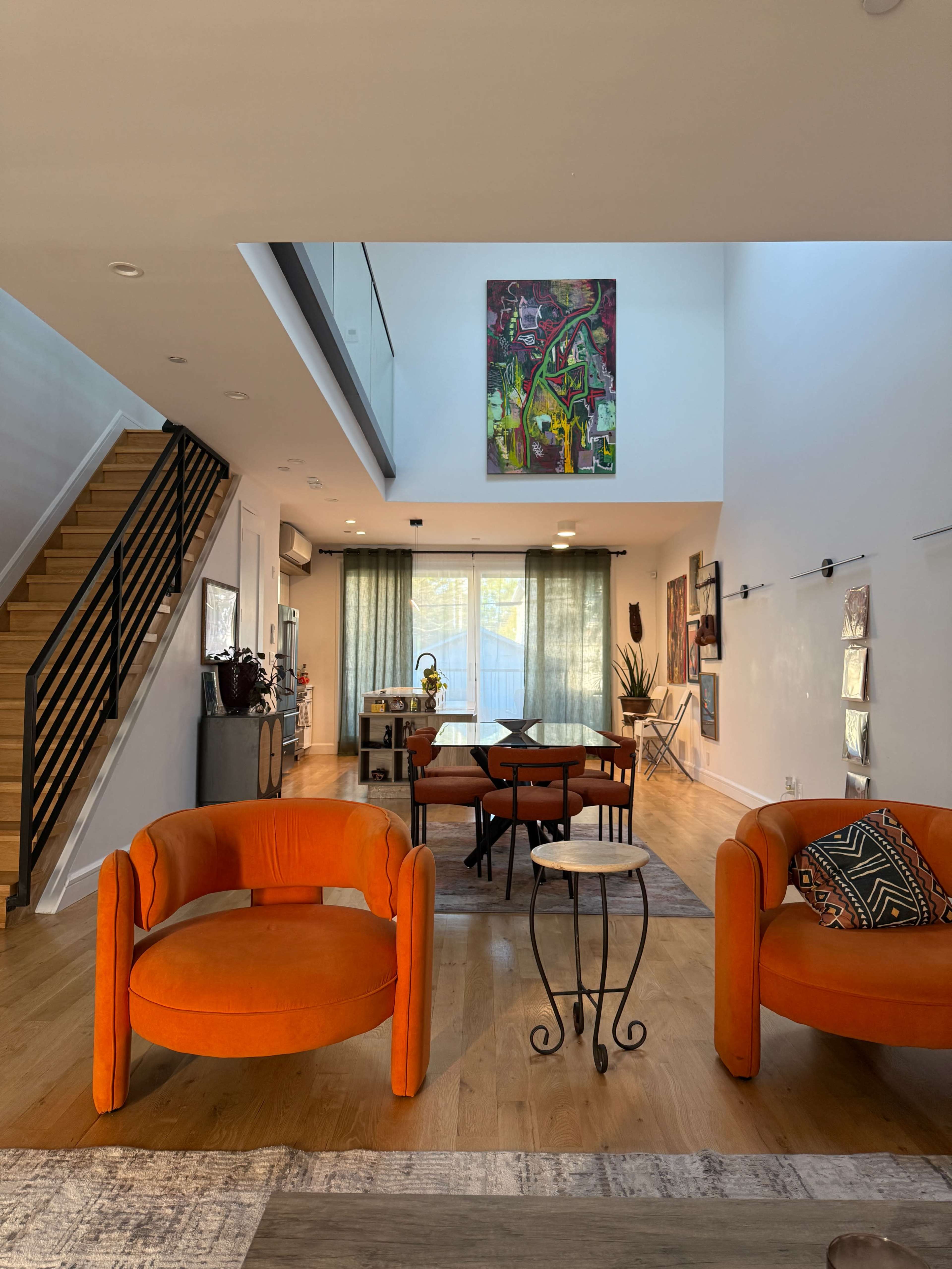 The image shows a spacious living area featuring orange circular chairs, a wooden dining table with chairs, and a staircase leading to an upper level, all illuminated by natural light from large windows.