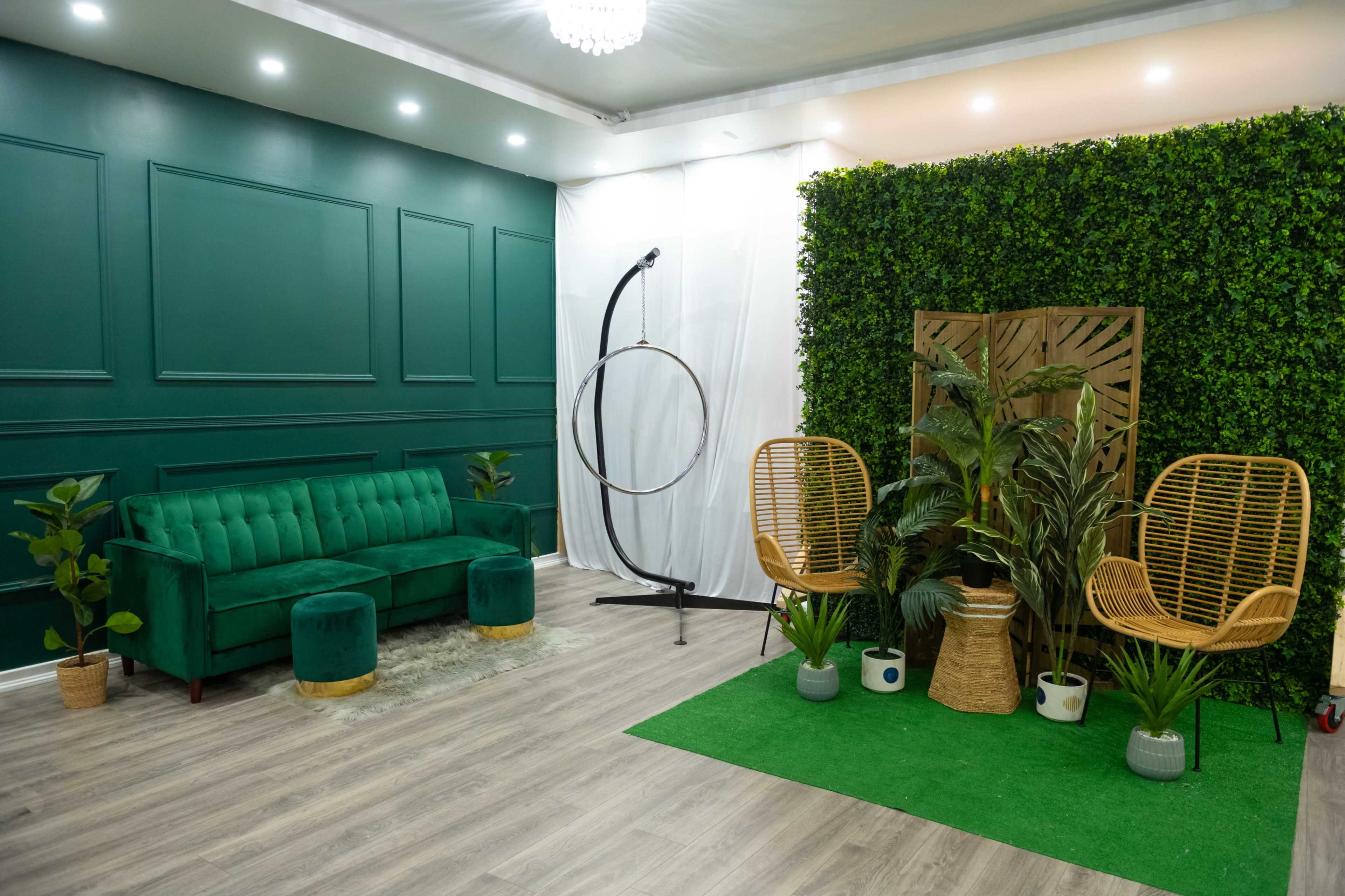 A modern indoor space featuring a green tufted sofa, two rattan chairs, a hoop structure, and decorative plants against a backdrop of artificial greenery.