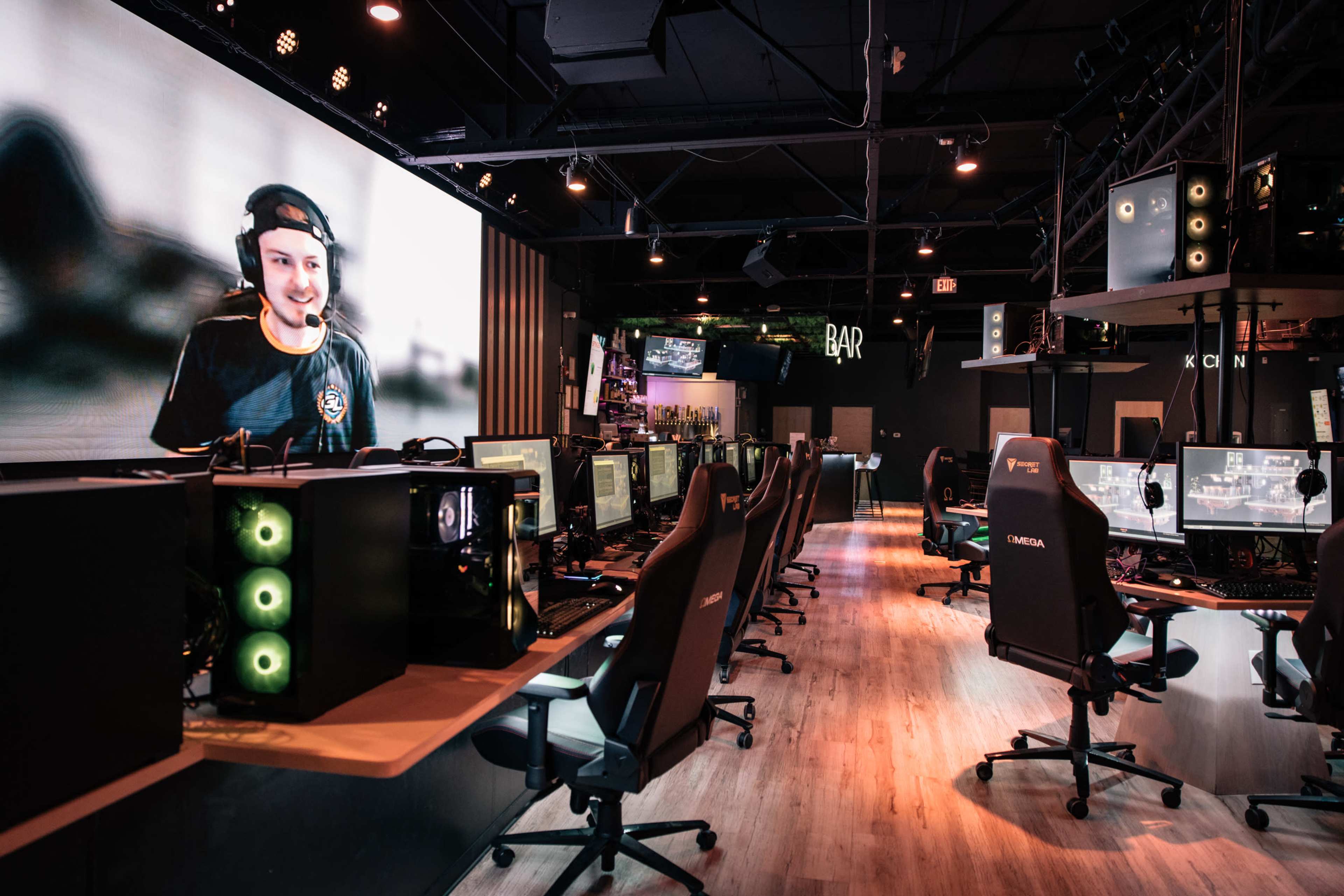 A gaming lounge with rows of computer gaming stations, a large screen displaying a player's face, and various gaming setups.