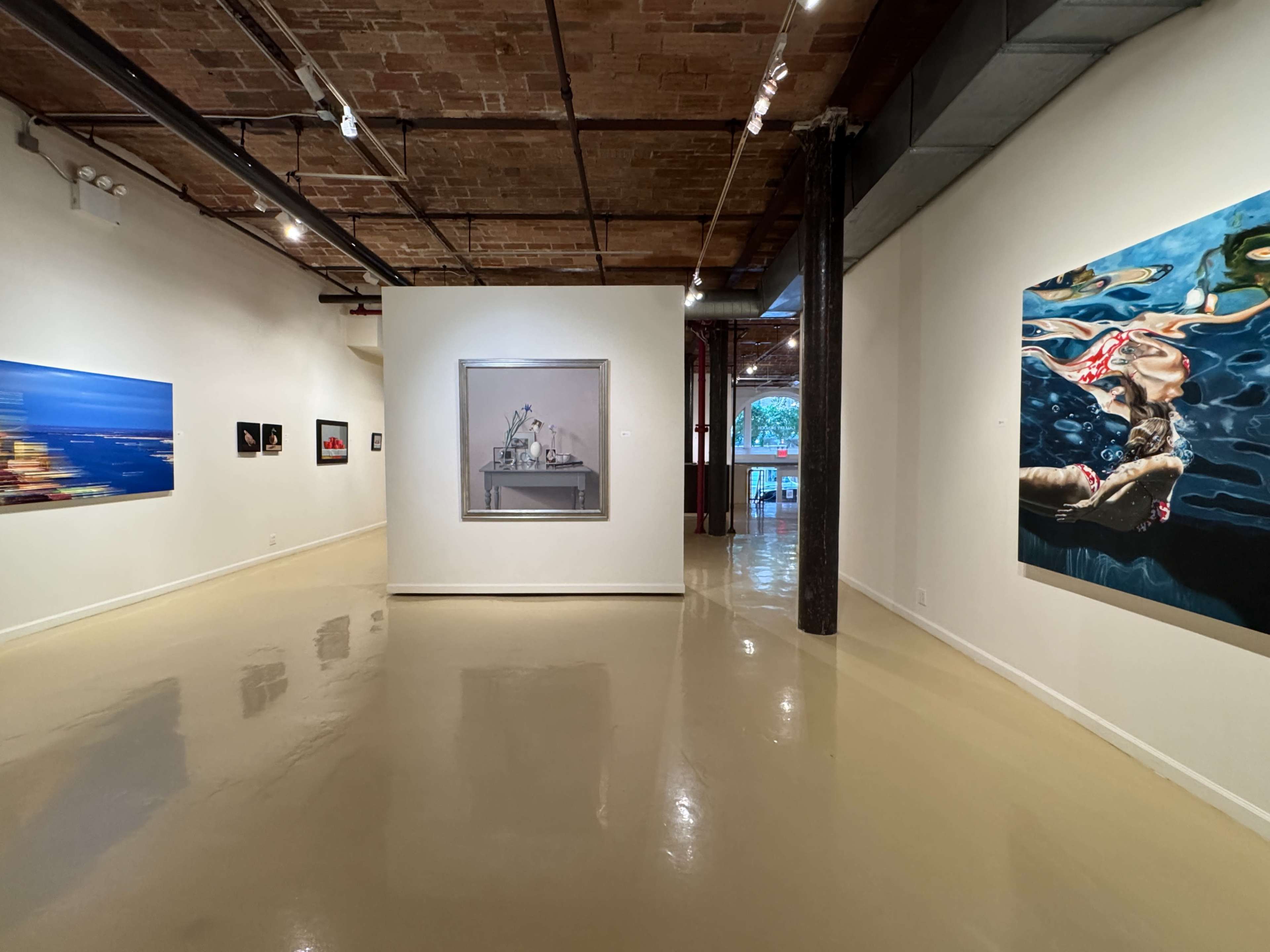 The image shows a contemporary art gallery with various artworks displayed on the walls and a polished floor.