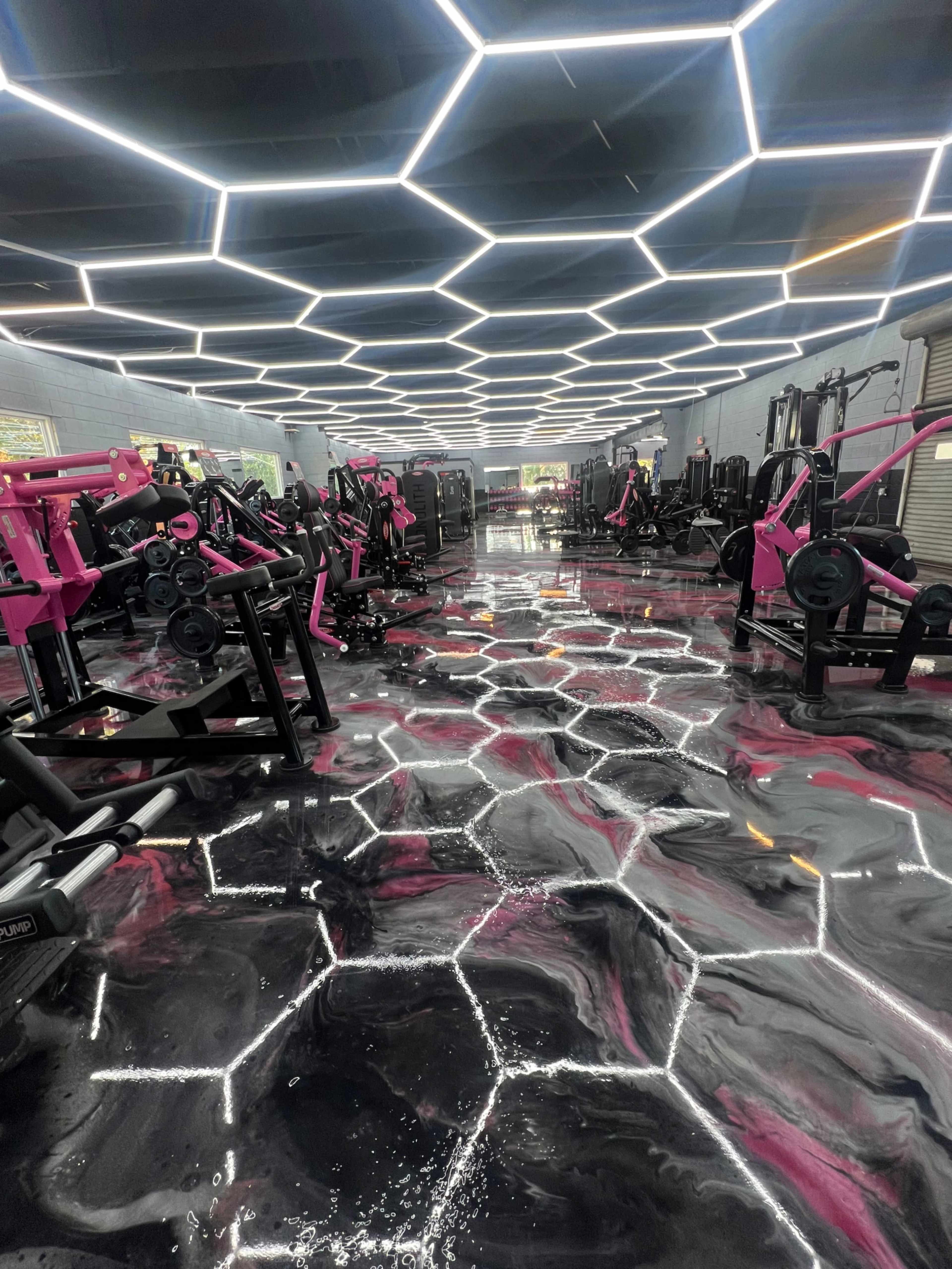 The image shows a modern gym featuring pink exercise machines and a glossy, hexagon-patterned floor illuminated by geometric lighting.