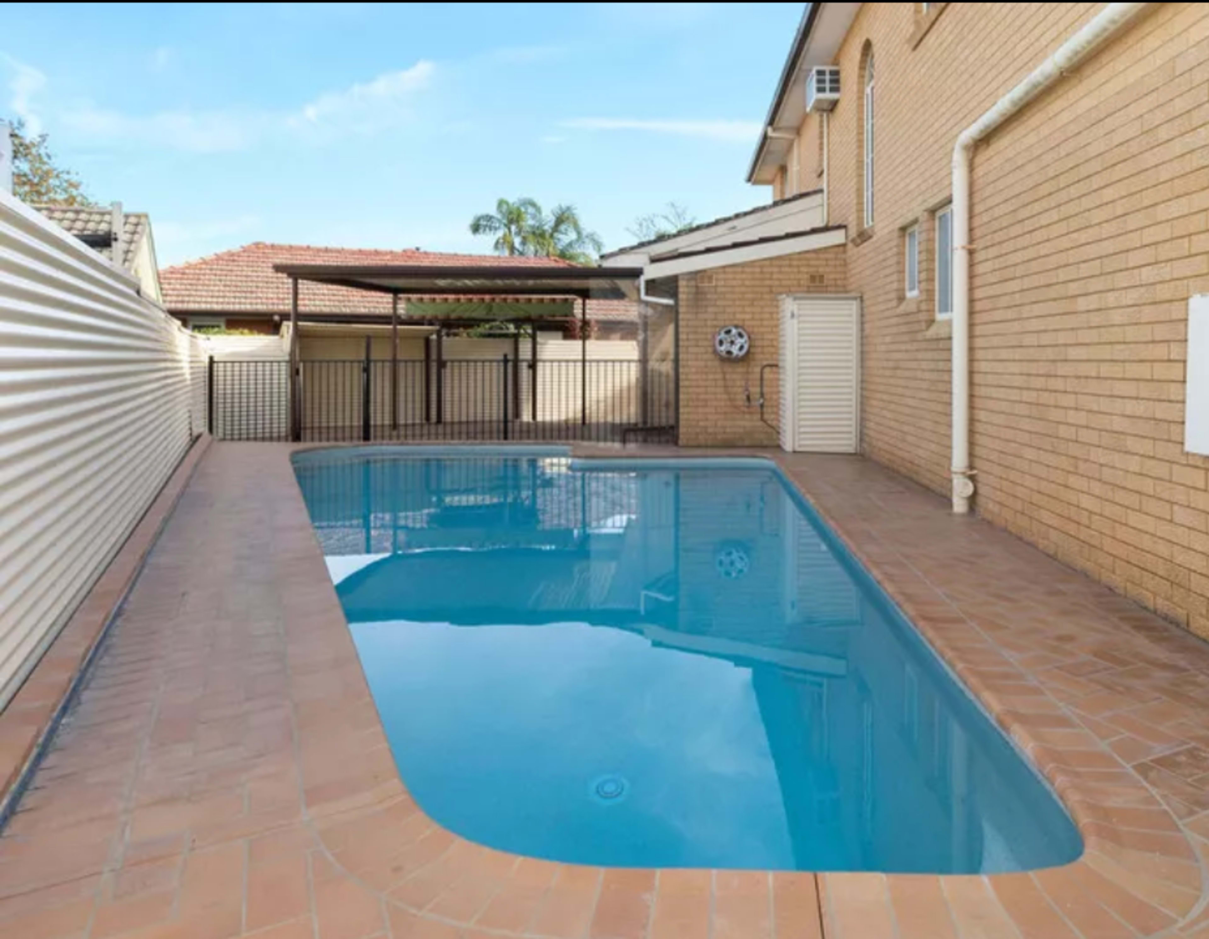 The image shows a rectangular swimming pool surrounded by tiled decking, with a wooden pergola in the background.