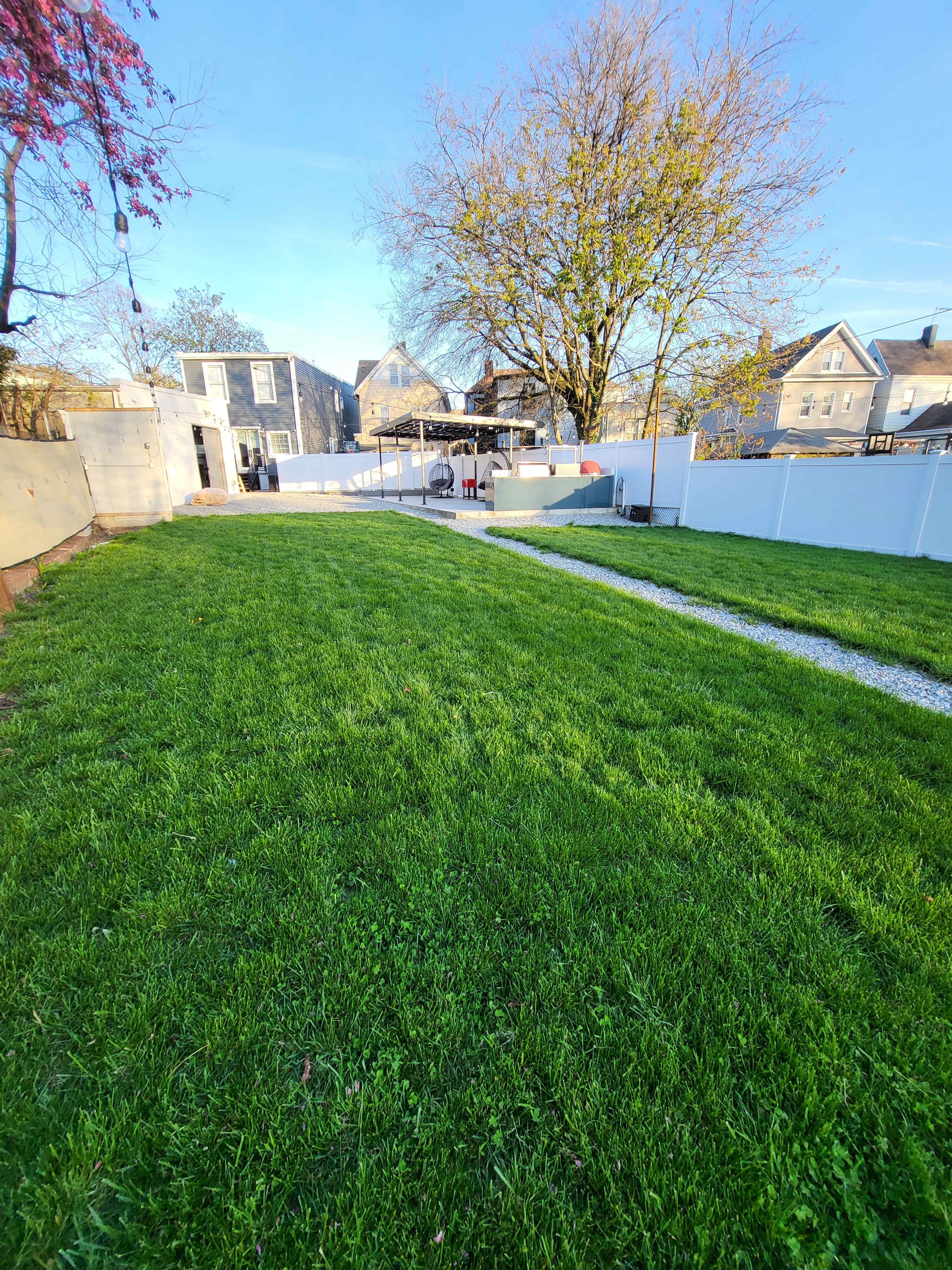The image shows a well-maintained green lawn leading to a fenced backyard with a patio area and several houses in the background.