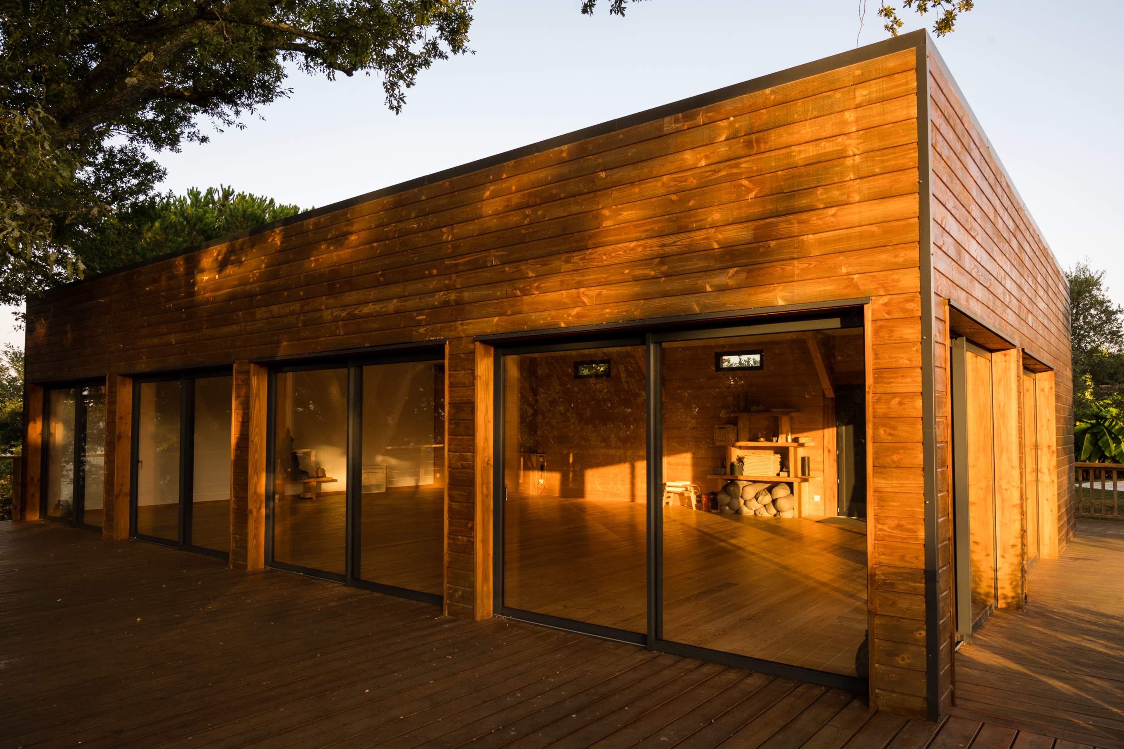 The image depicts a modern wooden building with large glass windows, situated on a wooden deck surrounded by greenery.