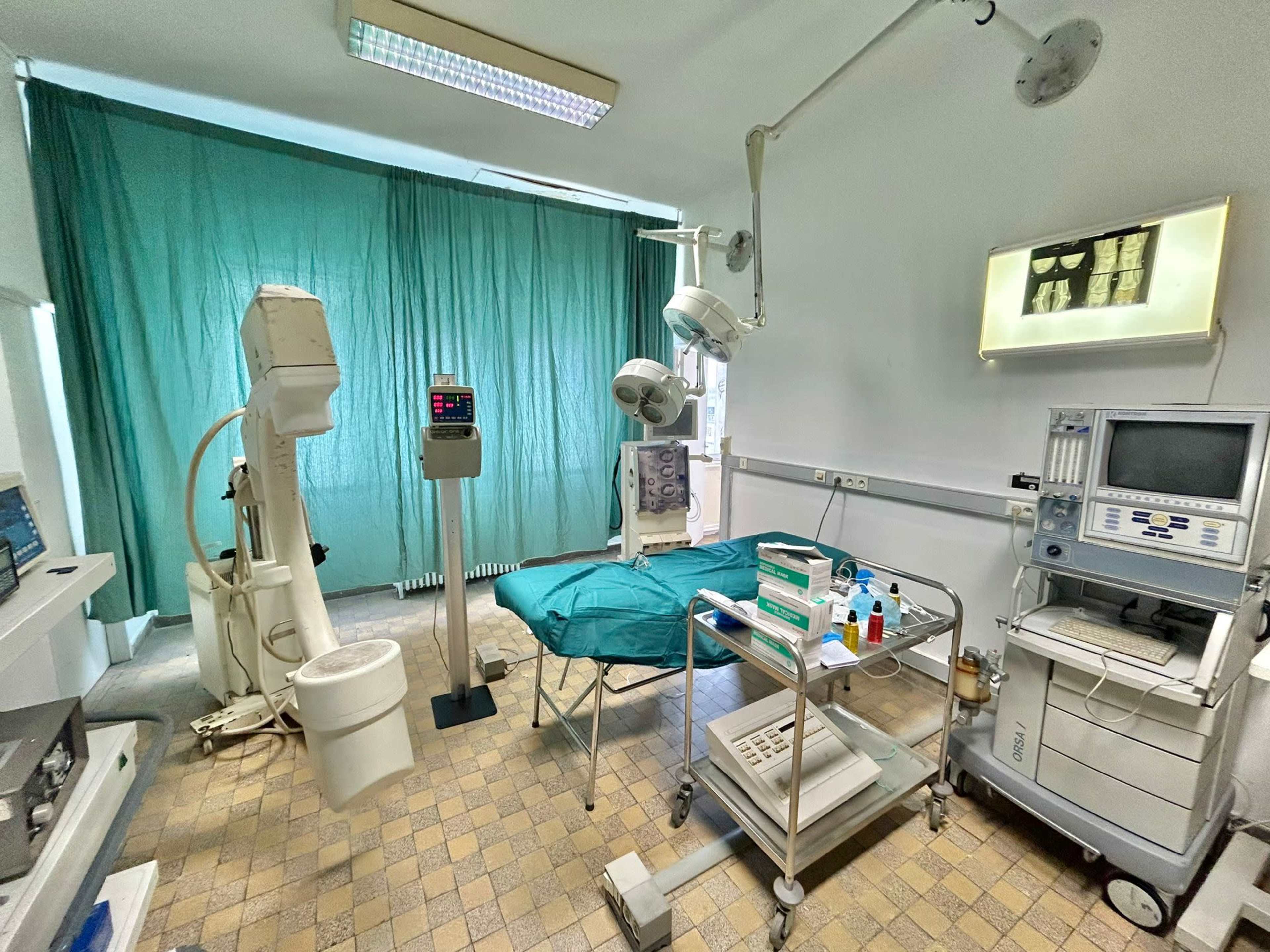 A medical examination room equipped with a surgical table, medical equipment, and a green curtain in the background.