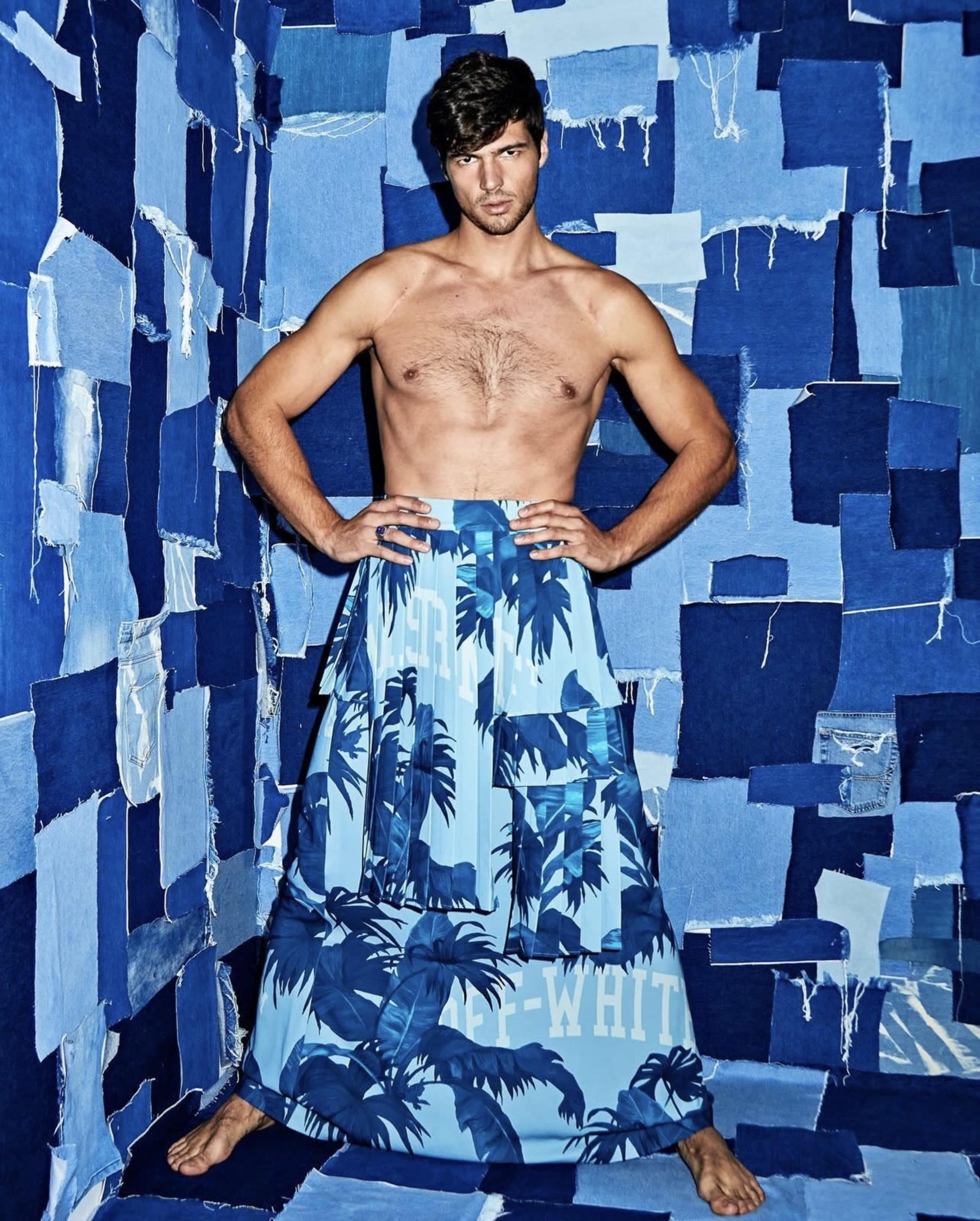 A shirtless man poses with his hands on his hips, wearing a long skirt made of blue and white patterned fabric, against a backdrop of blue denim patches.