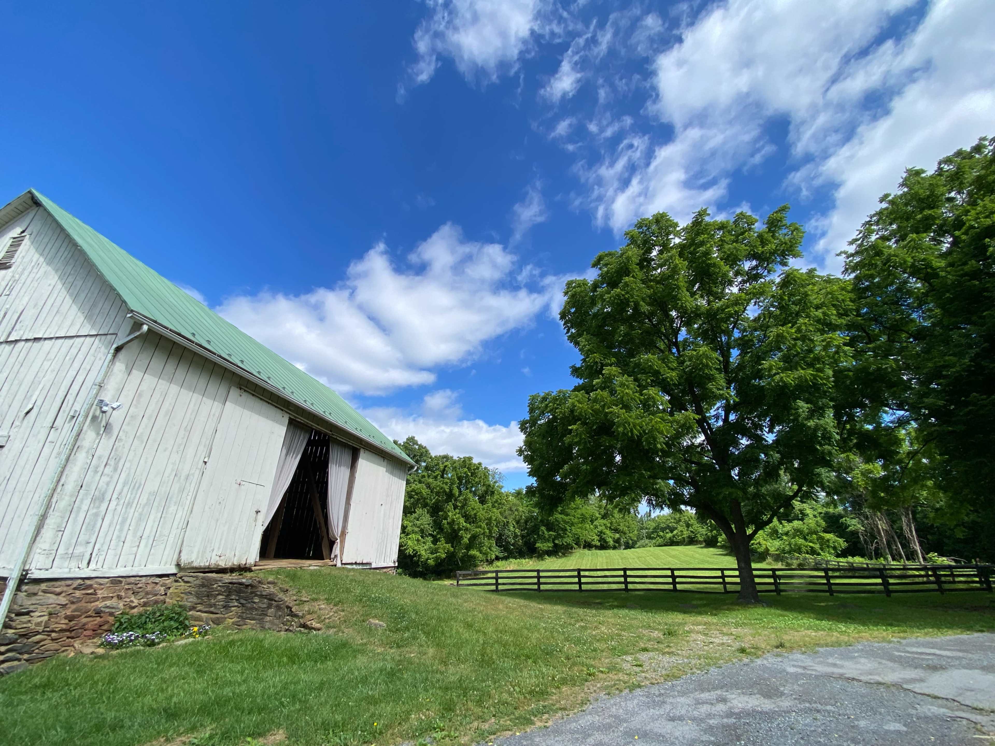 A white barn with a green roof stands beside a grassy area, with a large tree and a fenced field in the background under a partly cloudy sky.