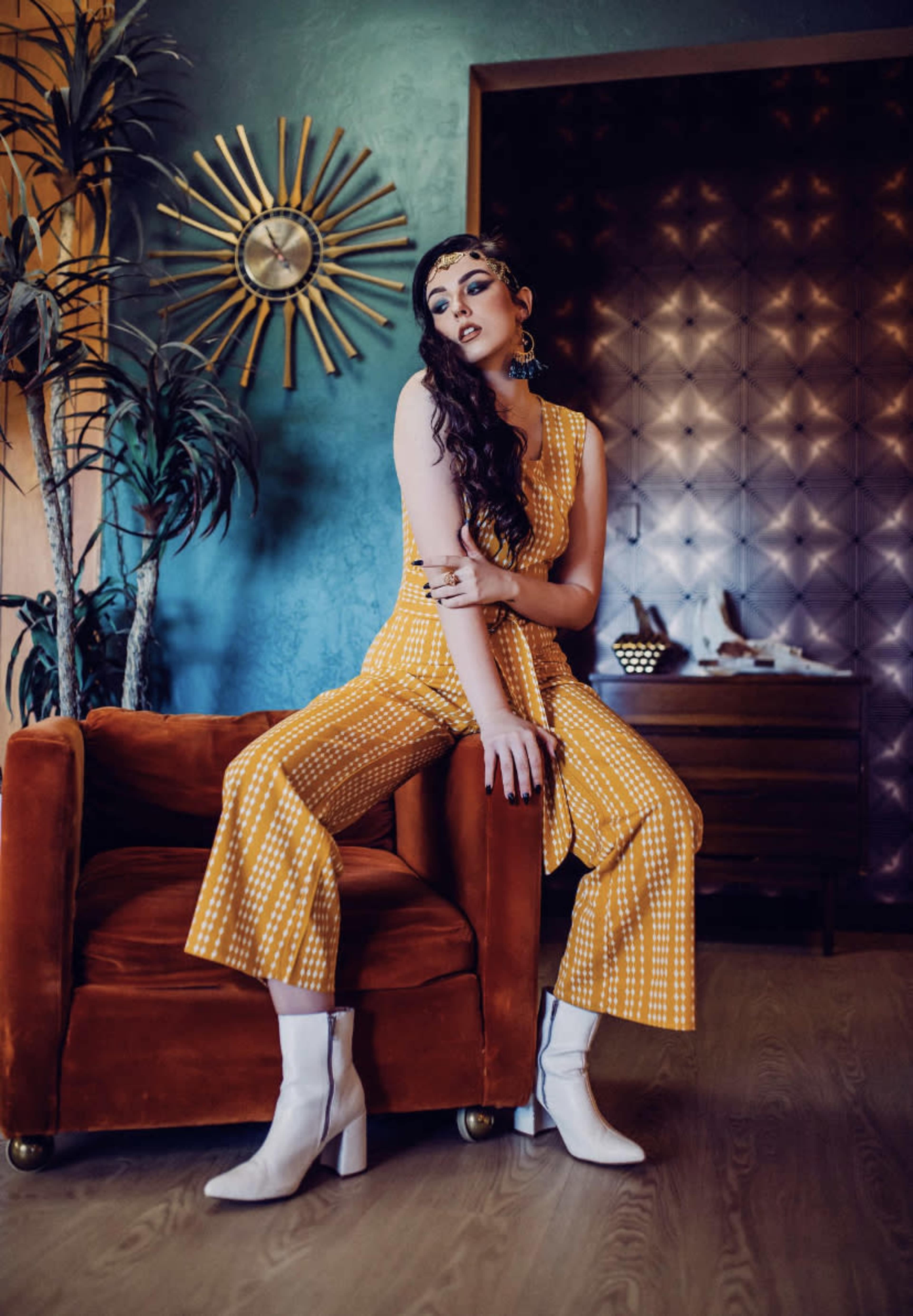 A woman wearing a yellow patterned jumpsuit and white boots sits on an orange velvet armchair in a stylish interior with mid-century decor.