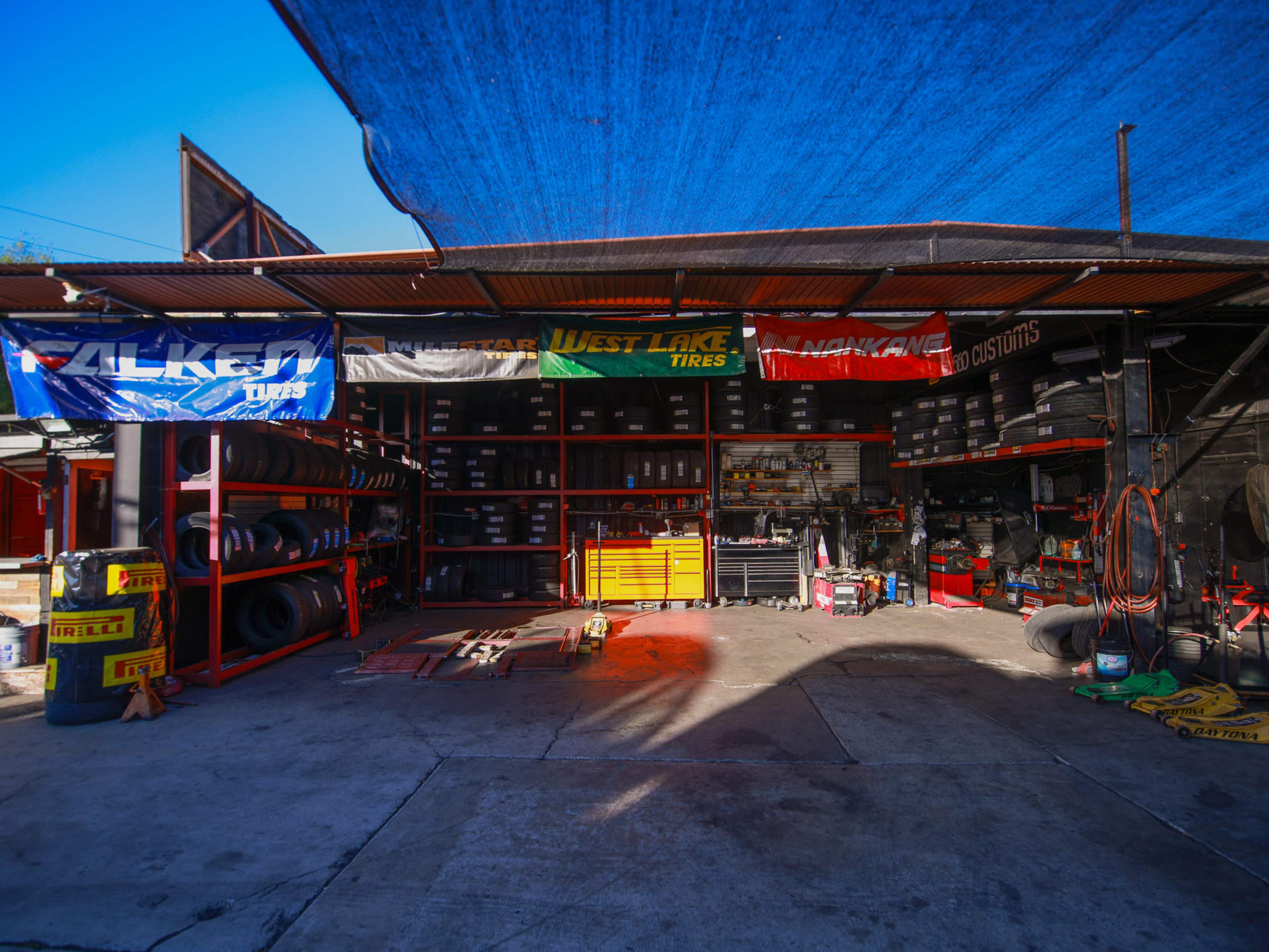 A tire shop features organized shelves stocked with tires, tools, and equipment under a blue canopy.