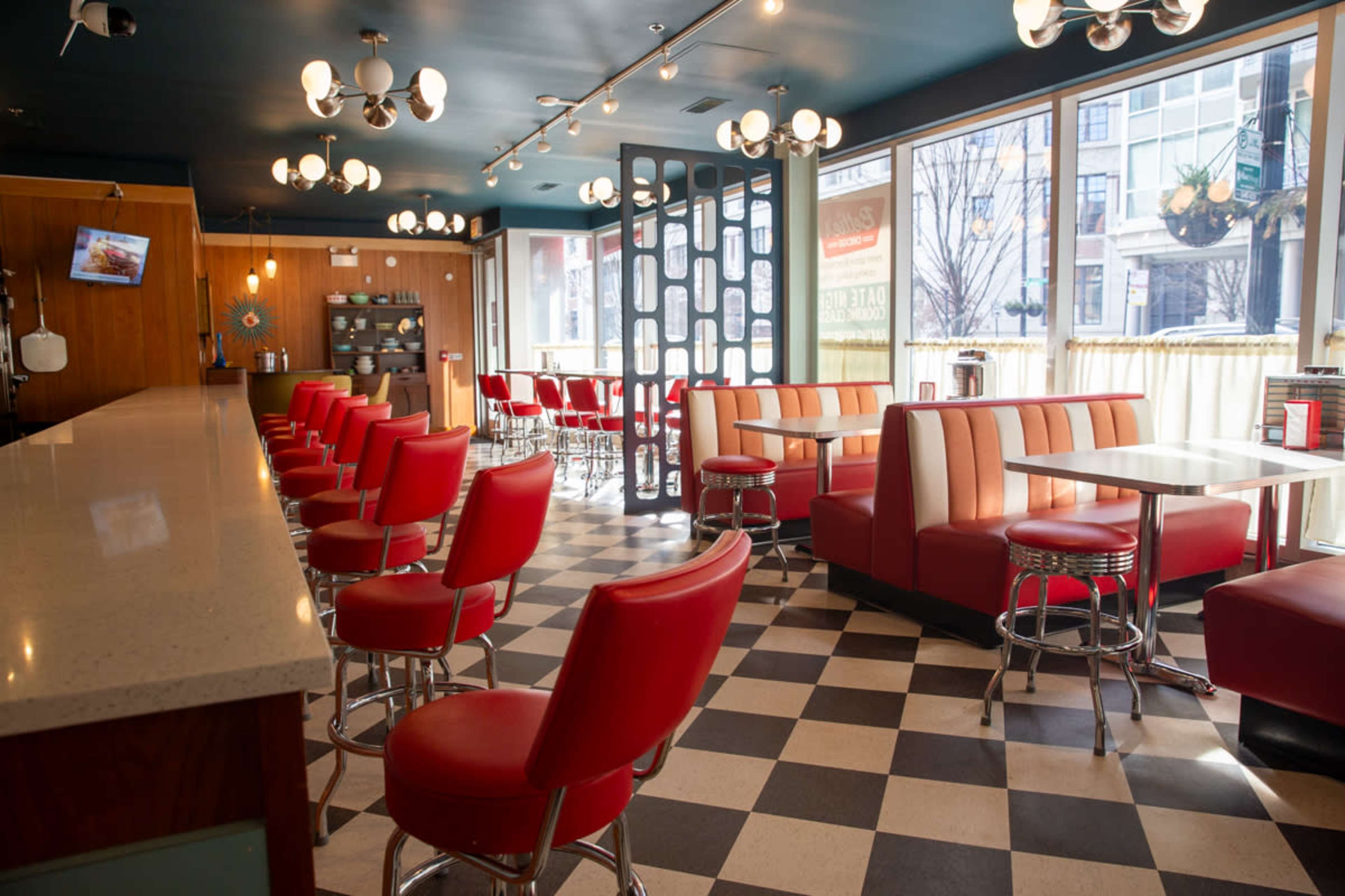 A retro diner features red upholstered chairs and a checkered floor, with a long counter and large windows overlooking the street.