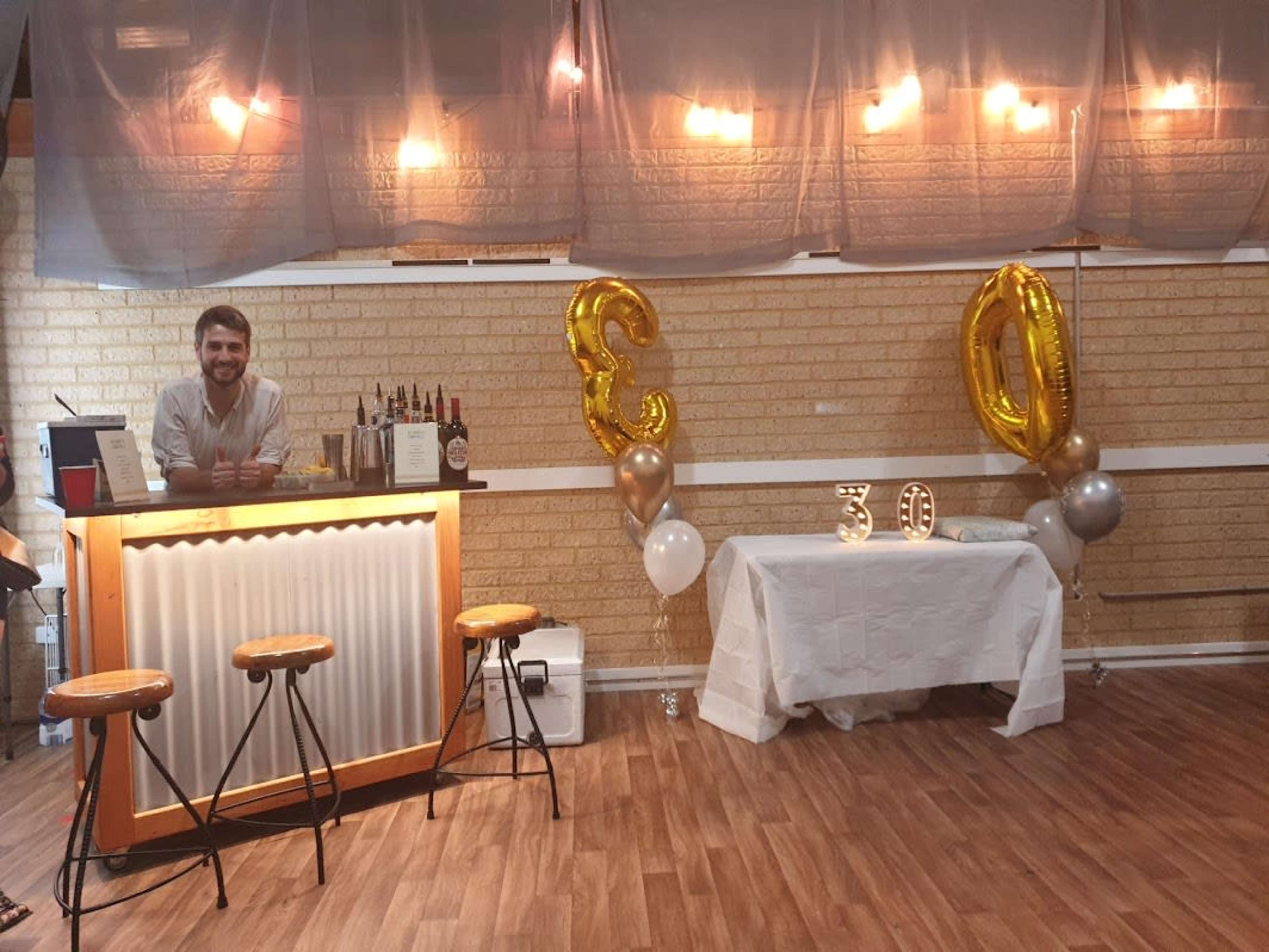 A bar setup is featured with a bartender behind the counter, a table decorated with balloons, and a sign displaying the number "30."