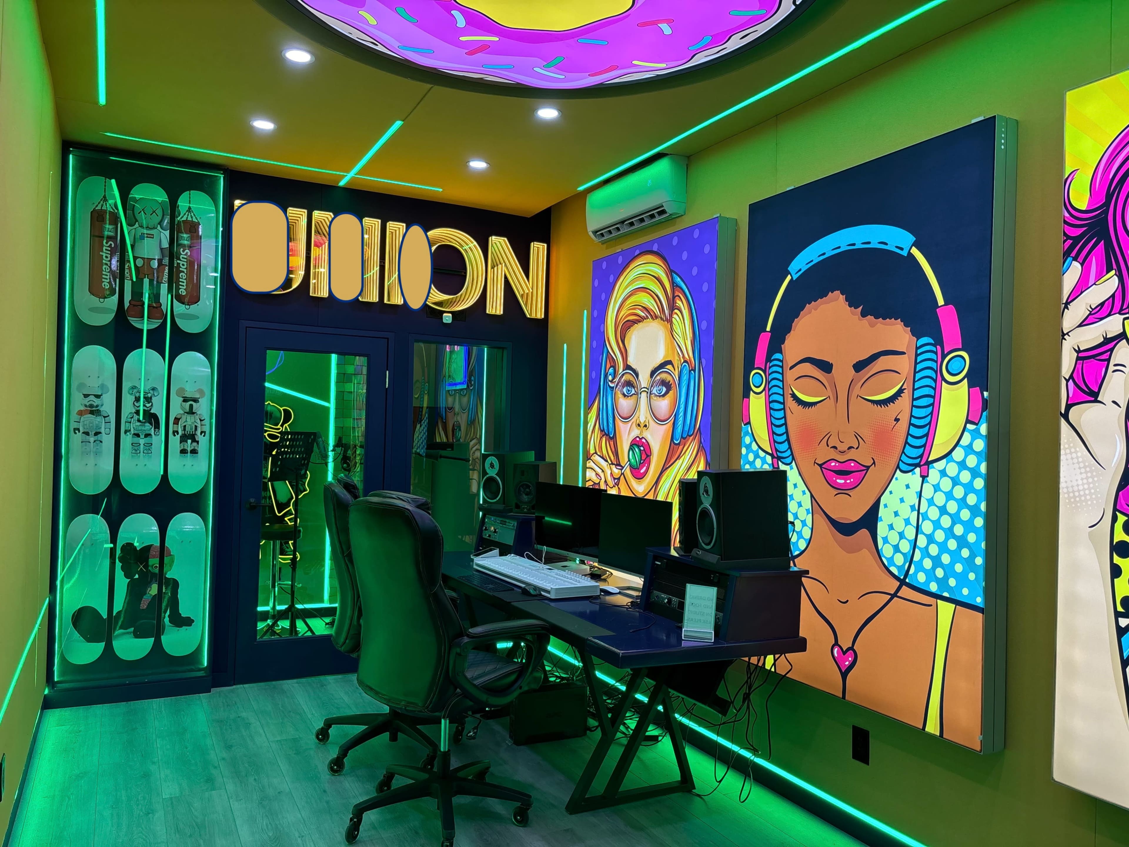 The image shows a vibrant studio space featuring colorful artwork on the walls, a desk with audio equipment, and illuminated accents in green and pink.
