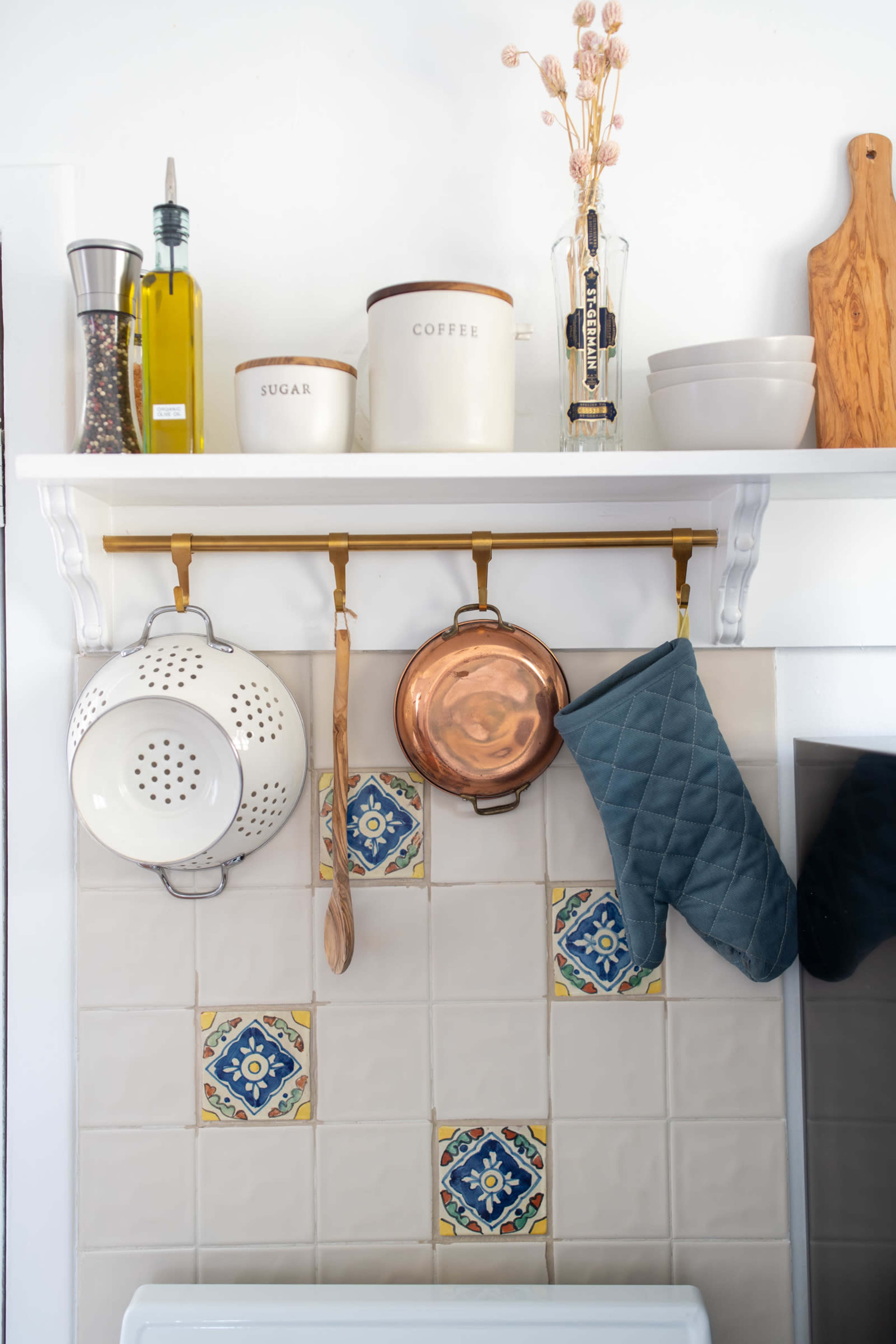 A shelf above a kitchen counter holds various containers and cooking utensils, including a colander, copper pot, and a blue oven mitt, with decorative tiles in the background.