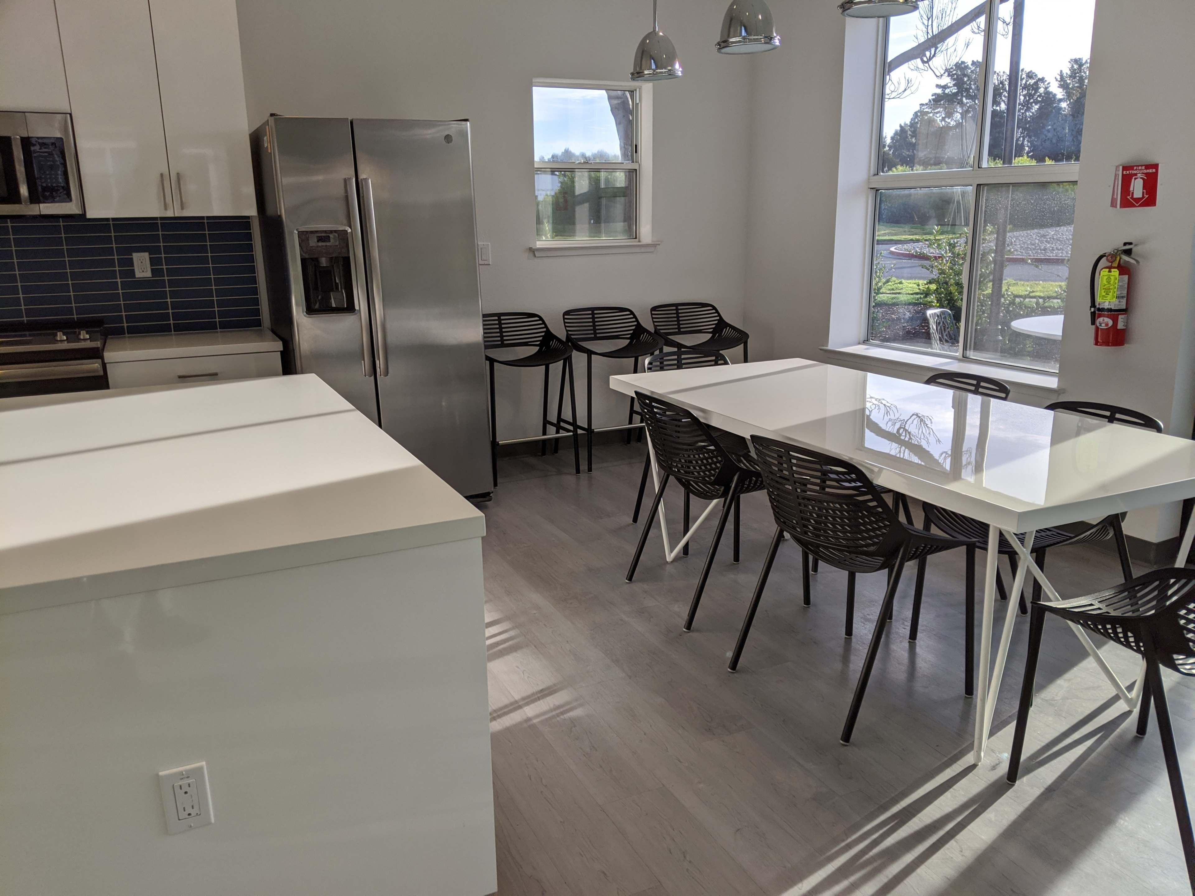 The image shows a modern kitchen and dining area with stainless steel appliances, a large white table surrounded by black chairs, and ample natural light coming through the windows.