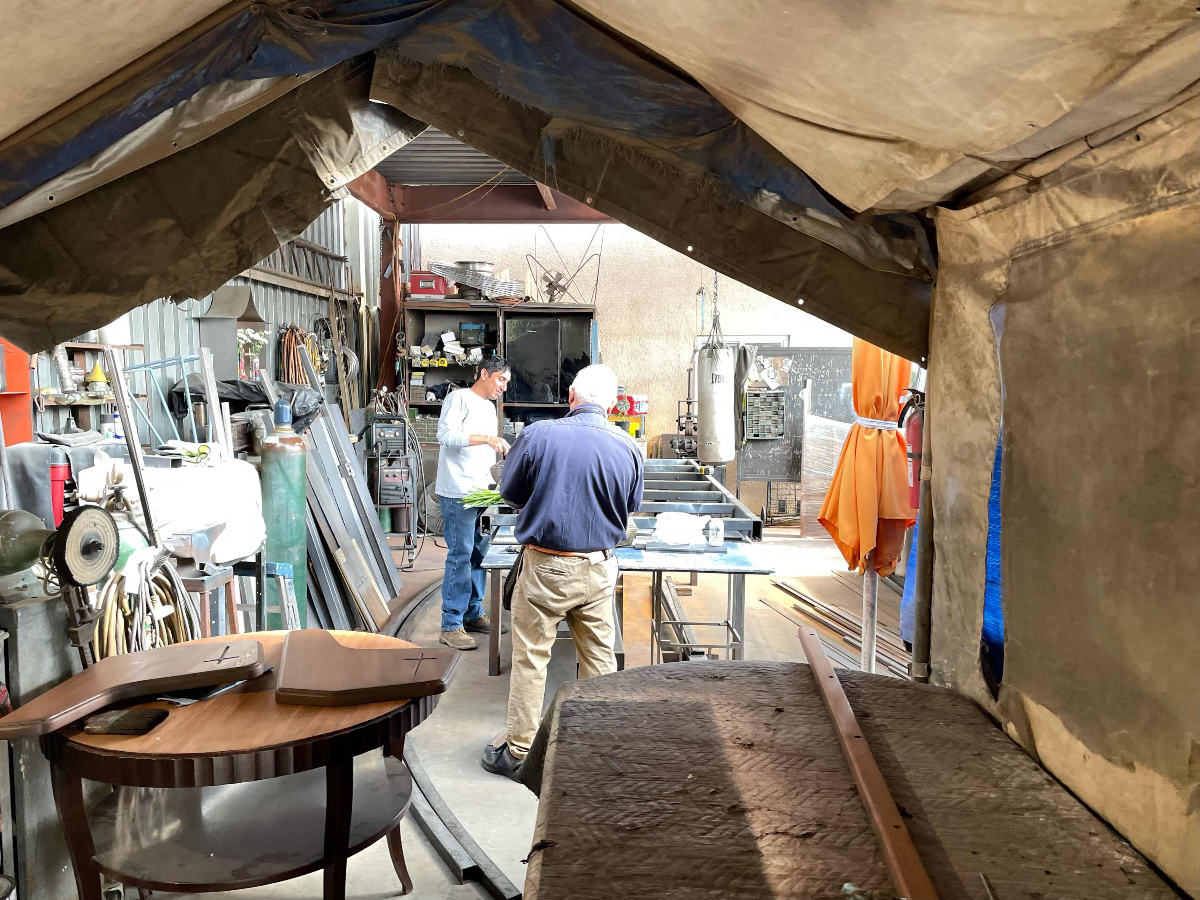Two men work at a table in a workshop filled with tools and materials, while covered by a tent-like structure.