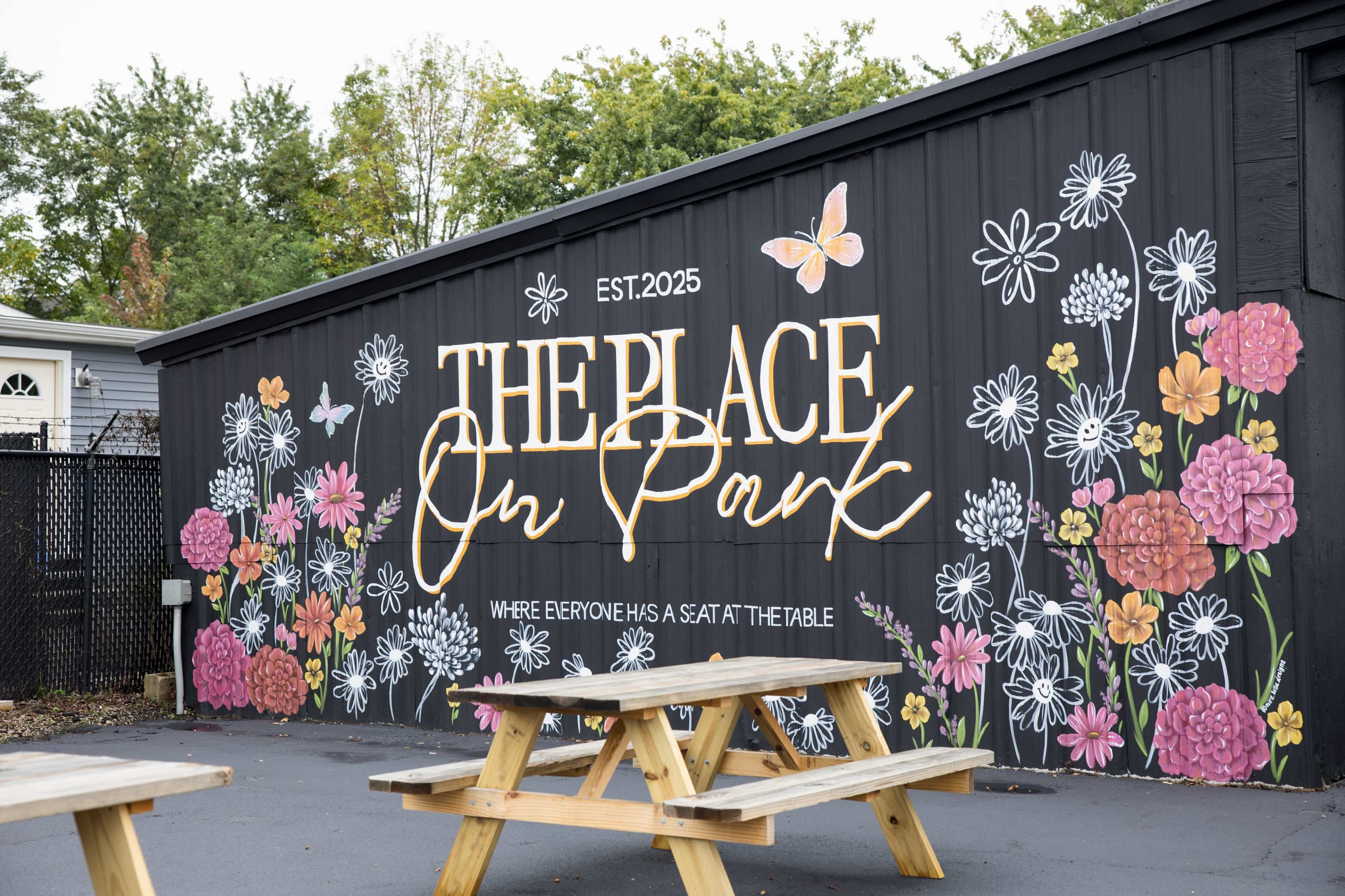 The image shows a colorful mural on a black wall that reads "THE PLACE On Park" with floral designs and a butterfly, along with the text "WHERE EVERYONE HAS A SEAT AT THE TABLE." A picnic table is situated in front of the mural.