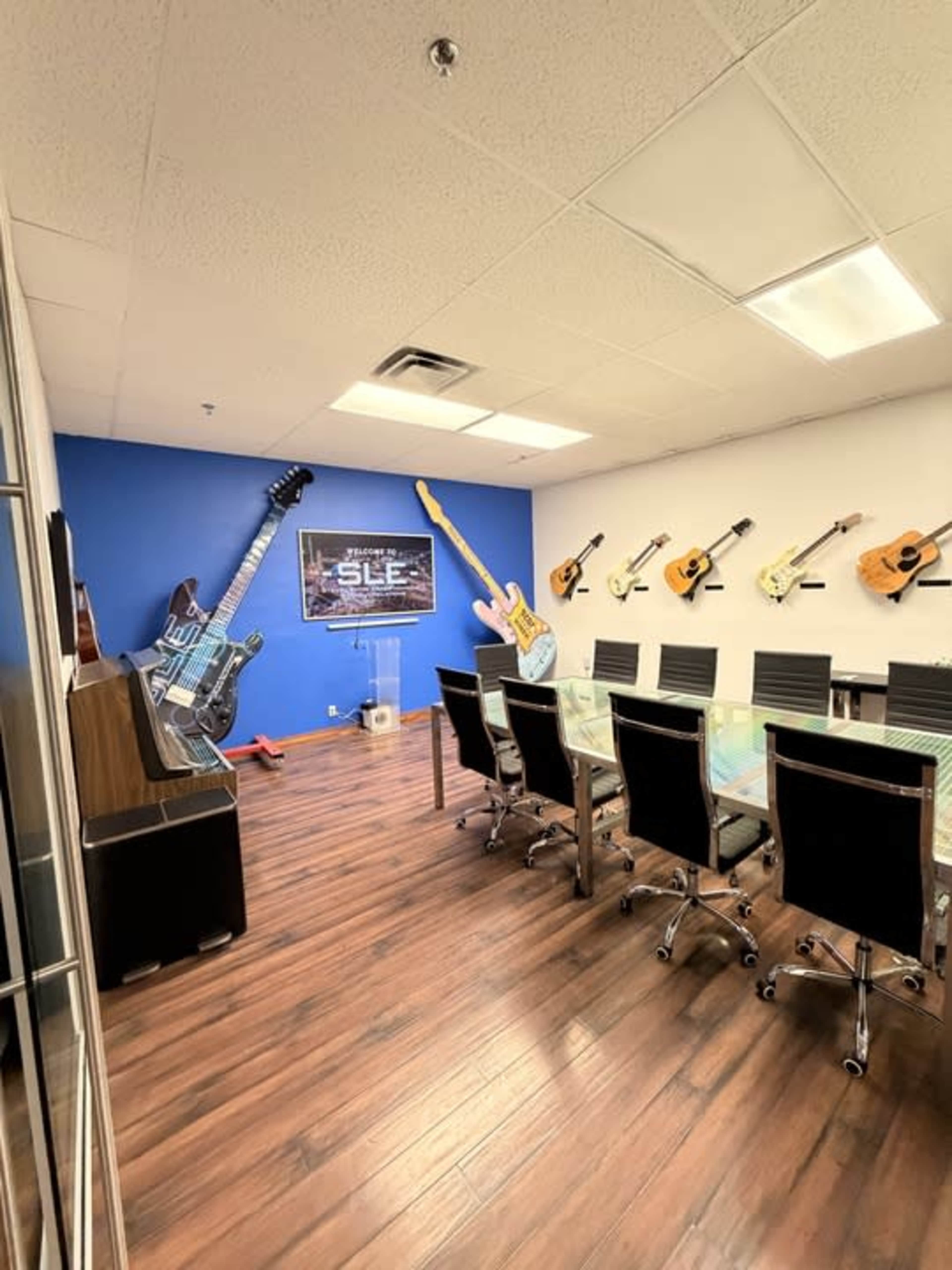The image shows a conference room featuring a long table surrounded by black chairs, with various guitars displayed on the blue wall and an upright piano in the corner.