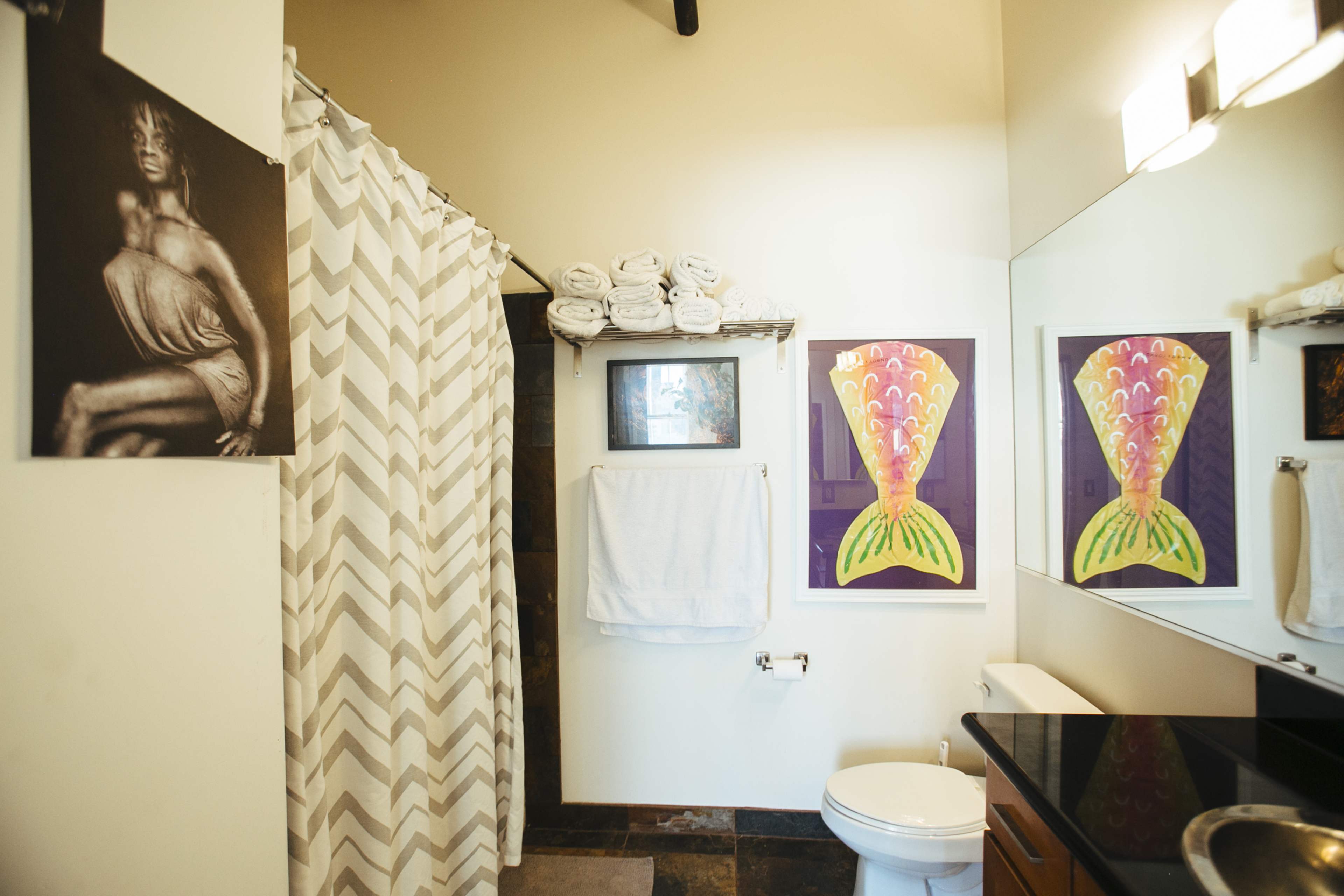 The image shows a bathroom featuring a shower curtain with a chevron pattern, neatly arranged towels on a shelf, two colorful framed art pieces, and a toilet adjacent to a sink with a dark countertop.