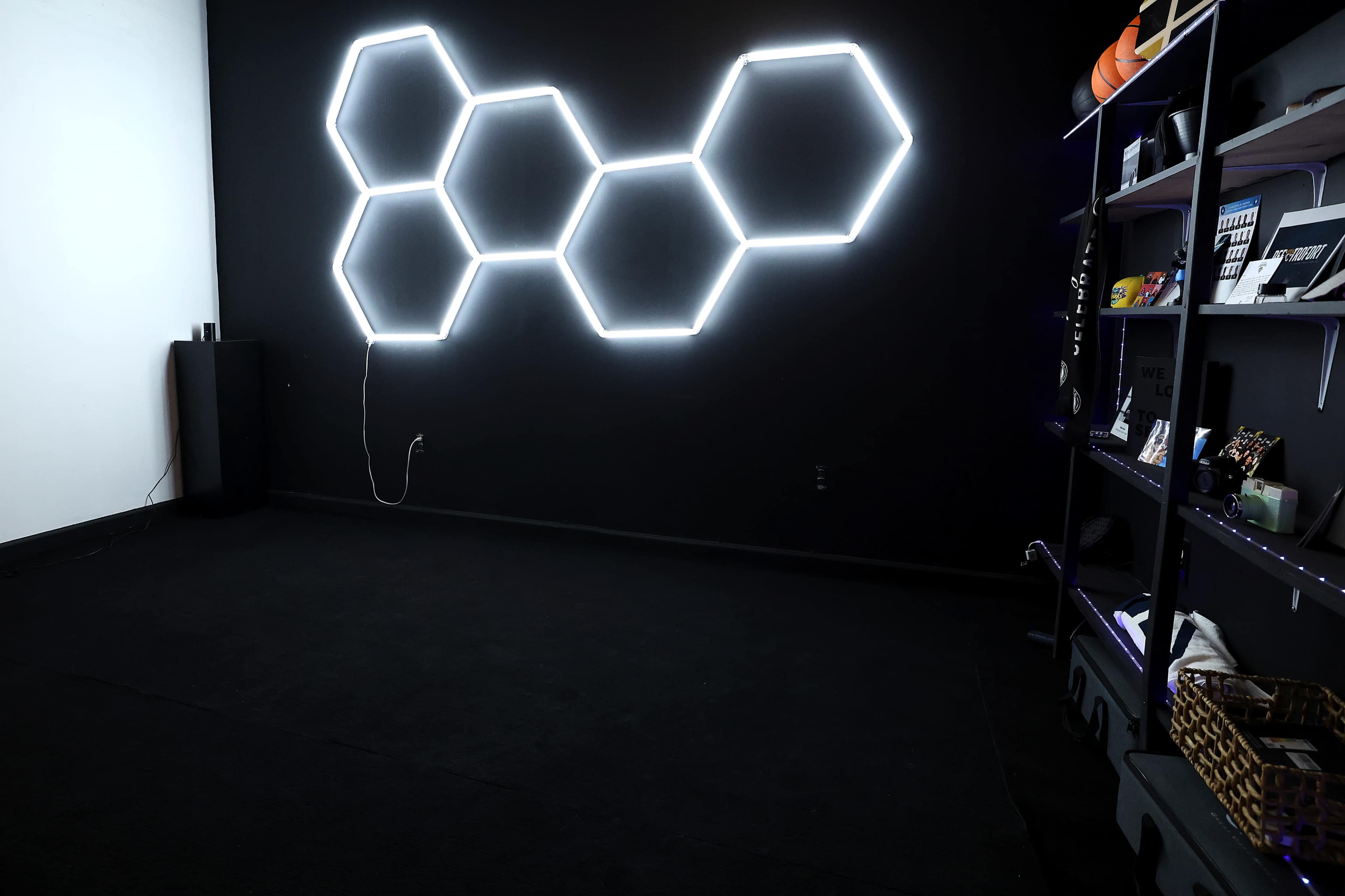 The image shows a room with a black wall featuring a hexagonal neon light installation, and a shelf on the right filled with various items.