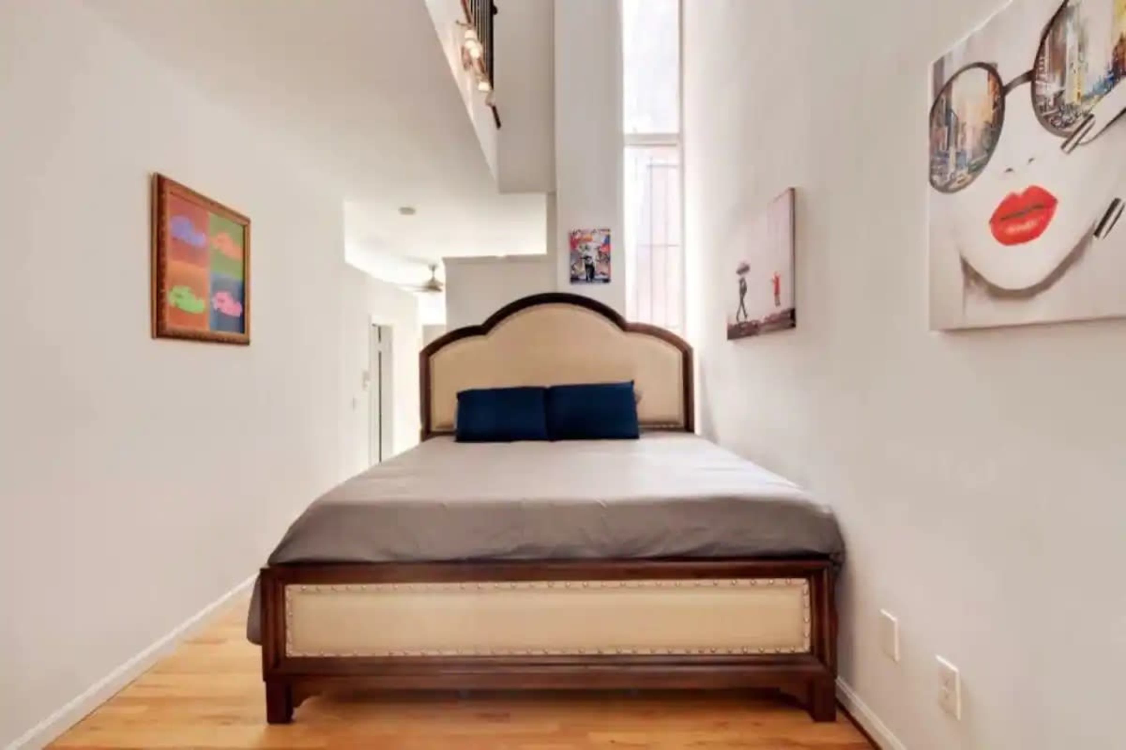 A narrow bedroom with a large bed featuring a decorative headboard, positioned along a white wall, and adorned with a couple of pieces of artwork.