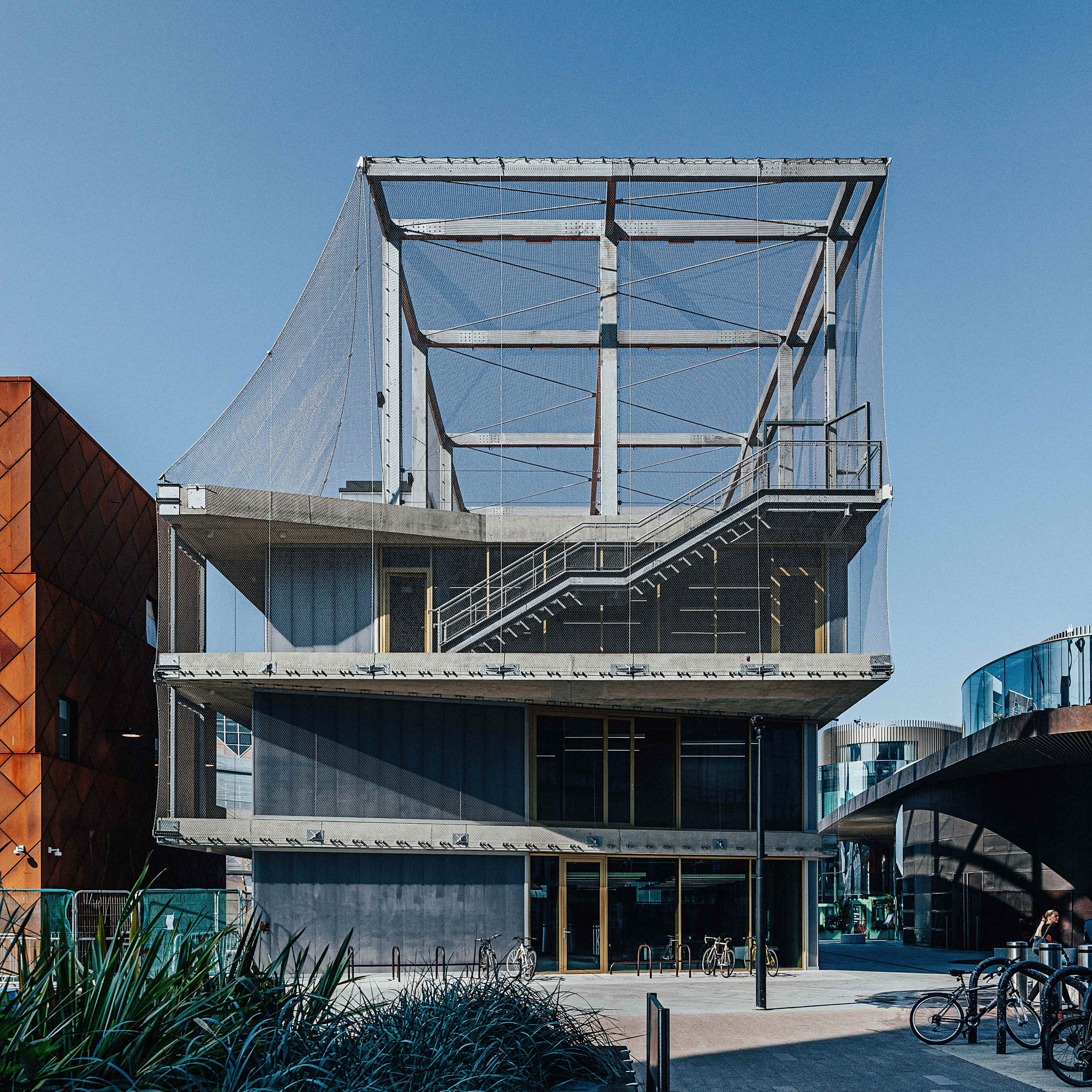 The image shows a contemporary three-story building with a distinctive angular design and a protective net covering the upper level.