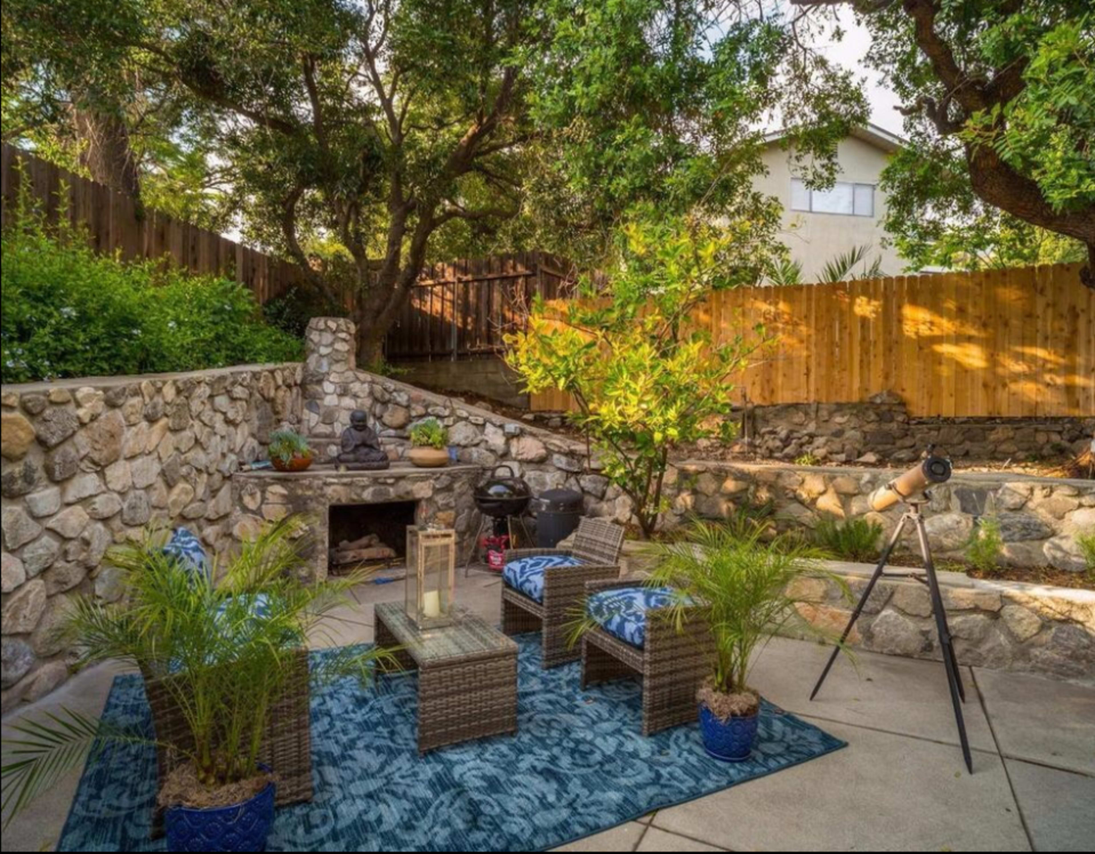 The image depicts a stone-walled outdoor patio with wicker chairs, a telescope, and potted plants surrounded by lush greenery.