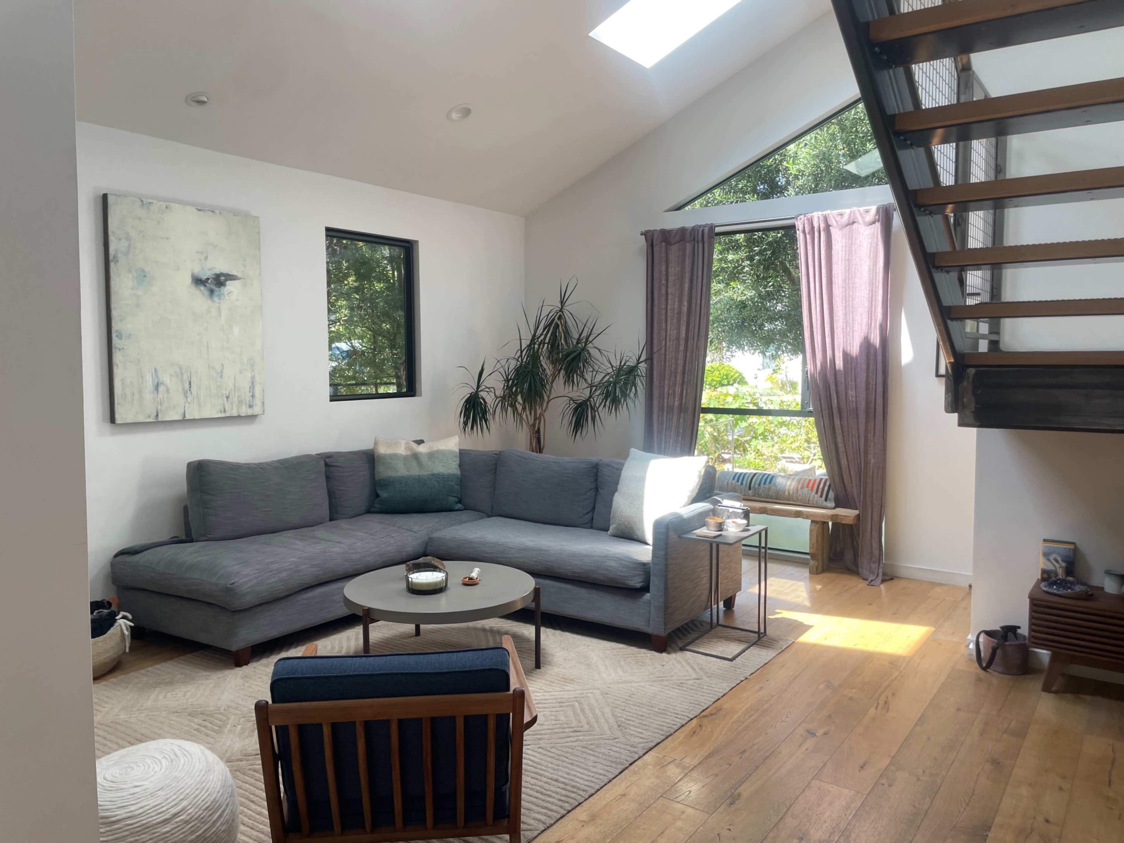 The image shows a bright living room with a gray sectional sofa, a round coffee table, and large windows overlooking greenery.