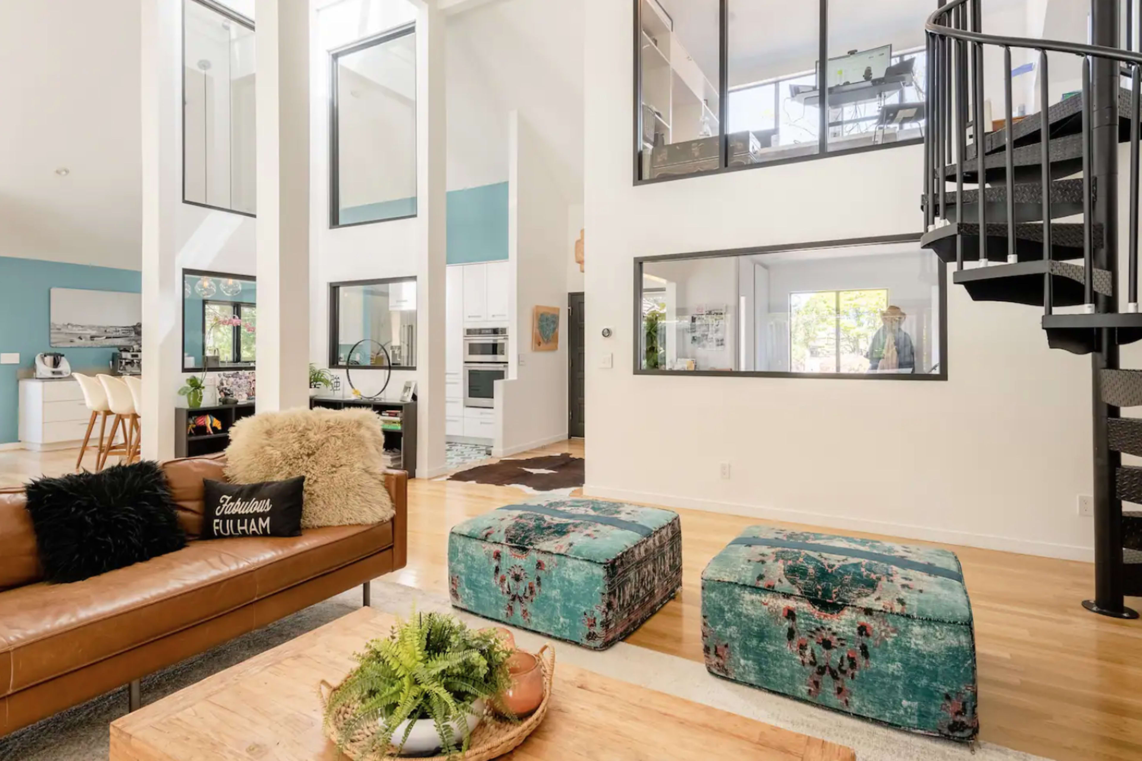 A modern living room with a brown leather sofa, two teal ottomans, and a spiral staircase leading to an upper level, featuring large windows and an open floor plan.