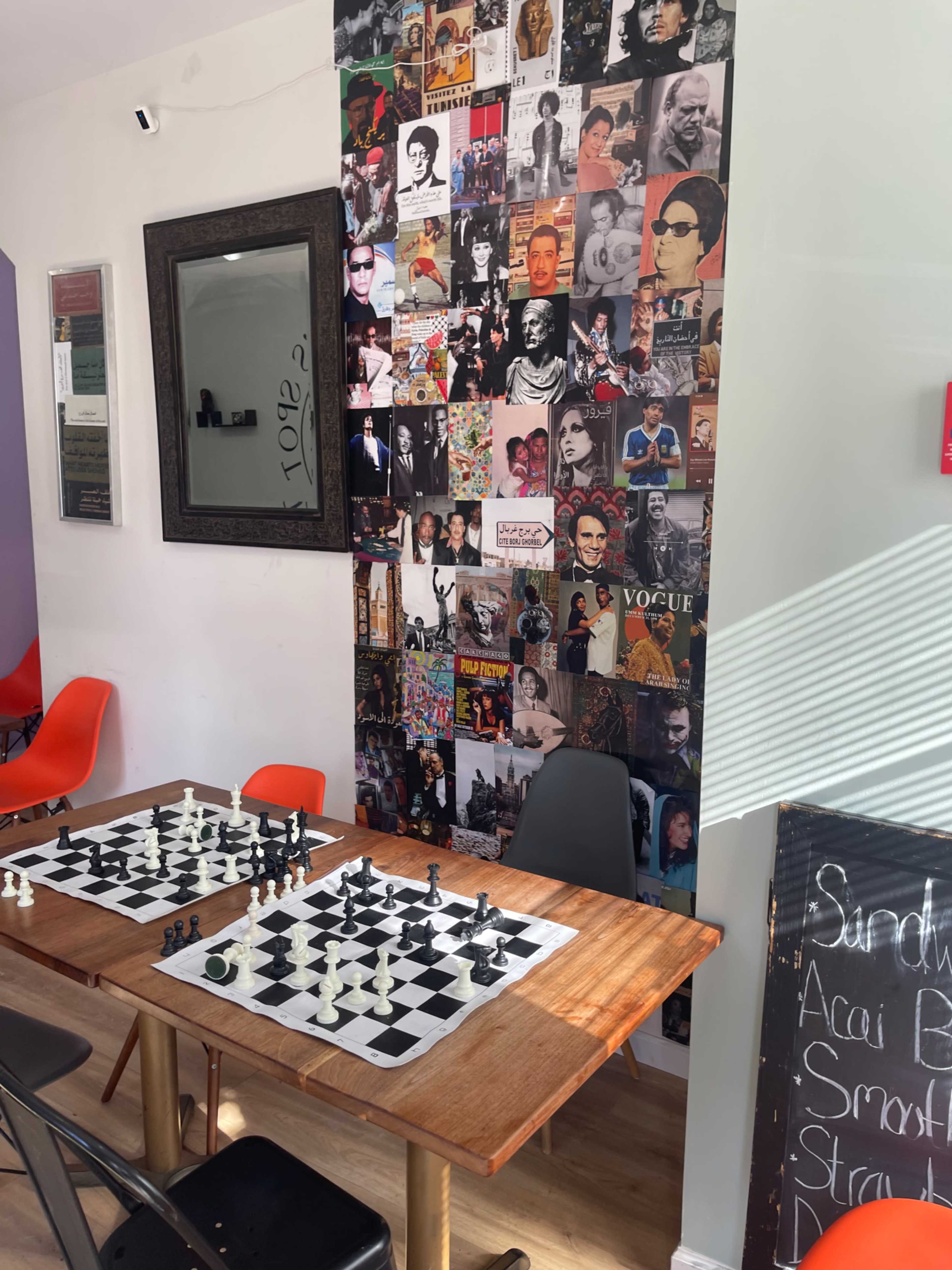 The image features a chess table with black and white pieces set up for a game, in front of a wall collage of various black and white photographs and posters.