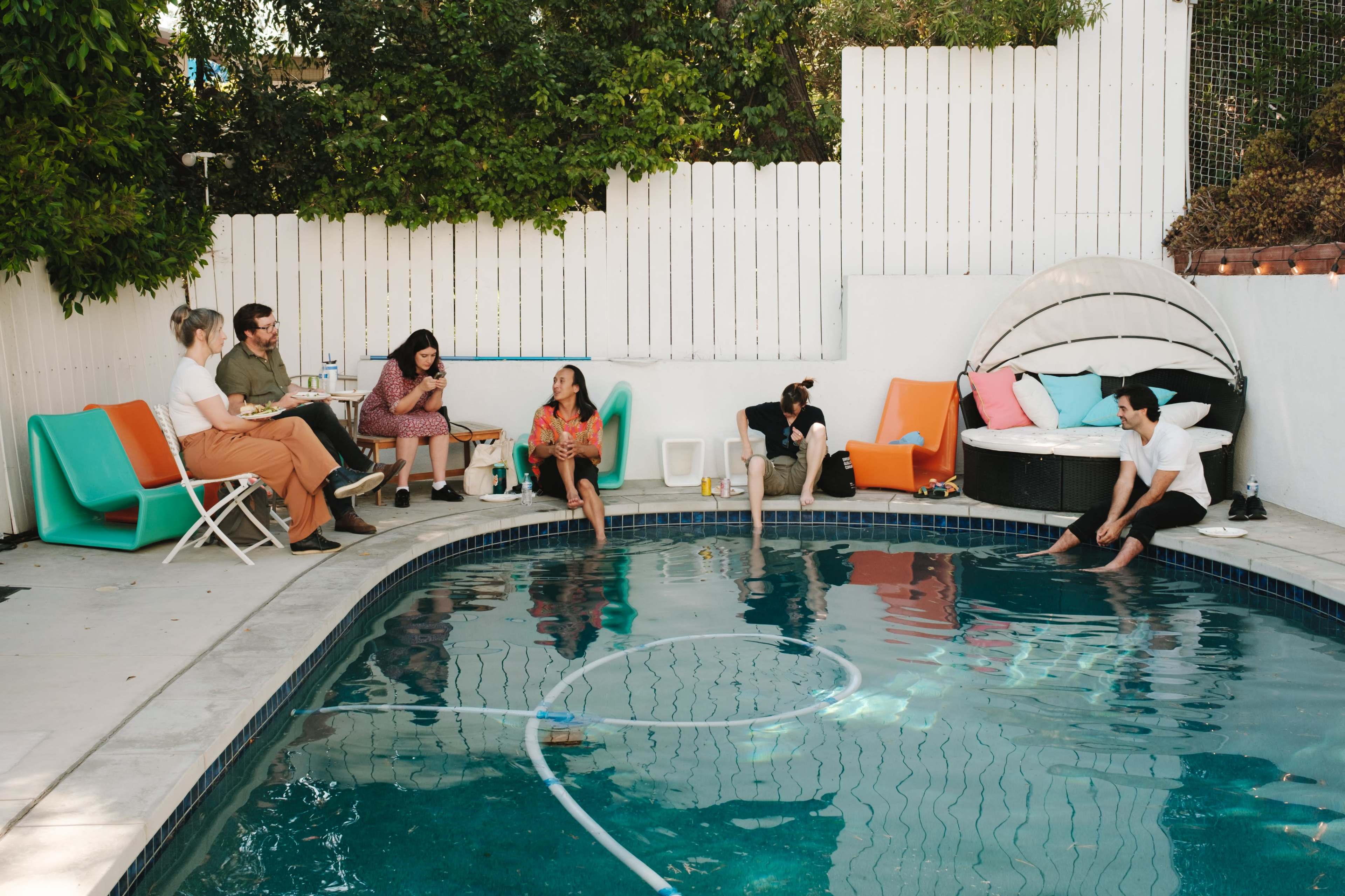 A group of six people is seated around a swimming pool in a backyard, with lounge chairs and a circular daybed nearby.