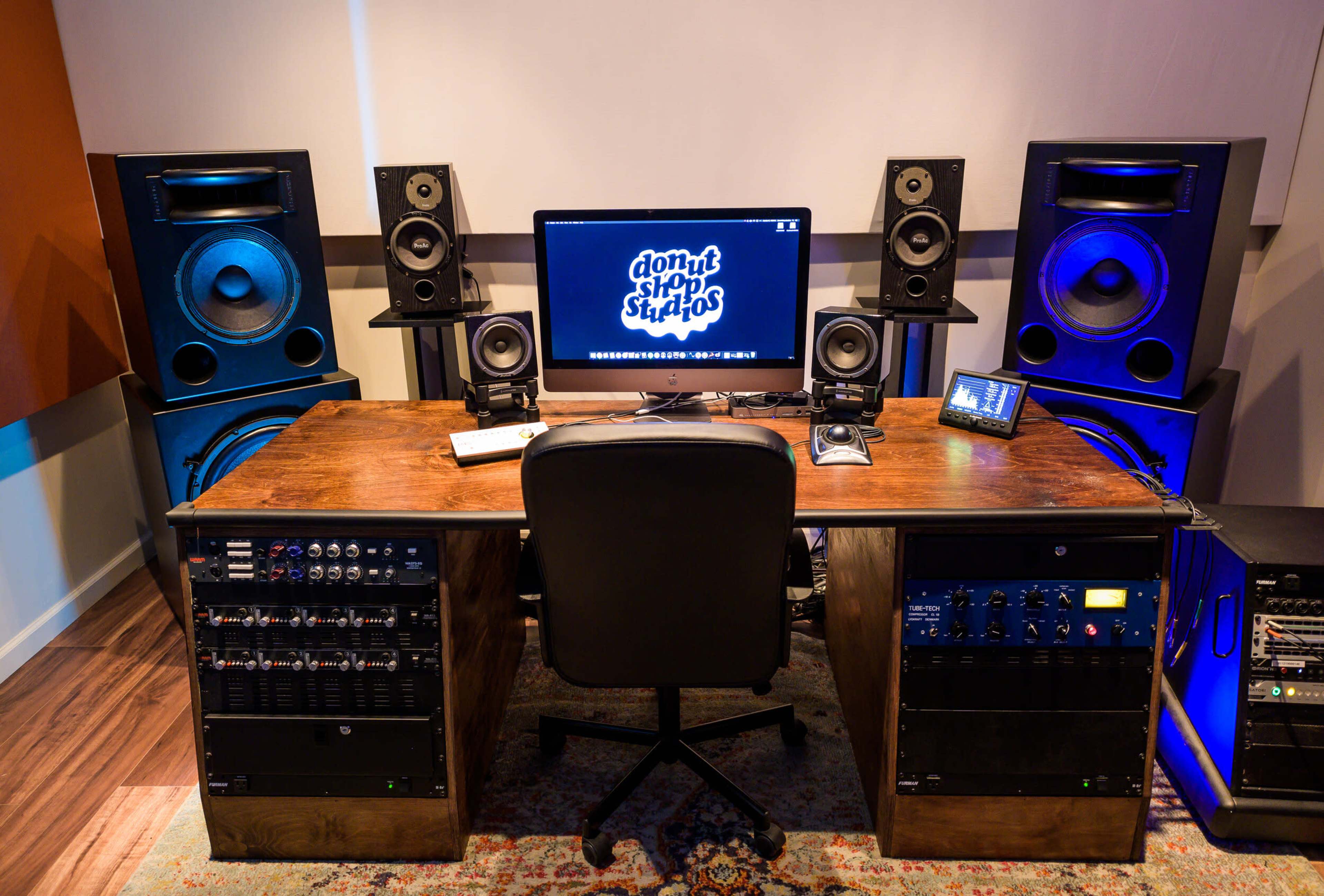 The image shows a music production studio with a desk equipped with a computer and audio equipment, flanked by large speakers on either side.