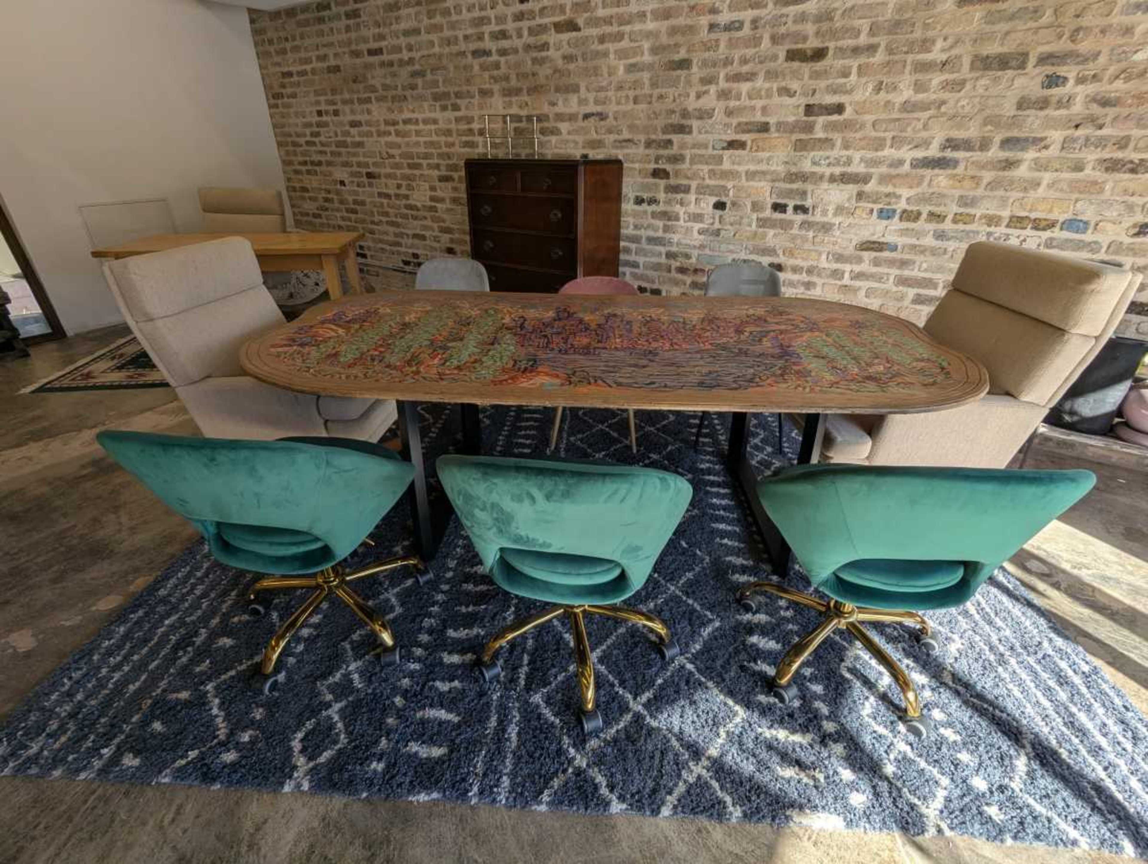 The image shows a large dining table with a colorful patterned top surrounded by four teal rolling chairs, set against a backdrop of exposed brick walls and hardwood flooring.