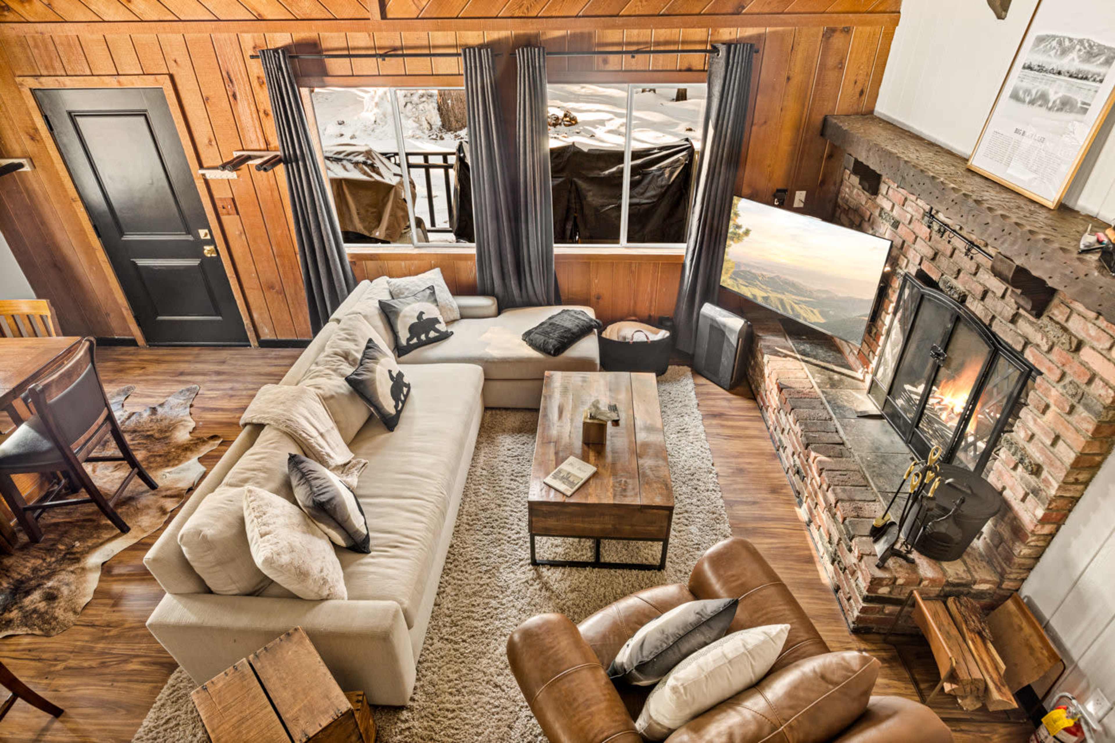 The image shows a cozy living room with a large sectional sofa, a wooden coffee table, and a brick fireplace, all illuminated by natural light from a window.