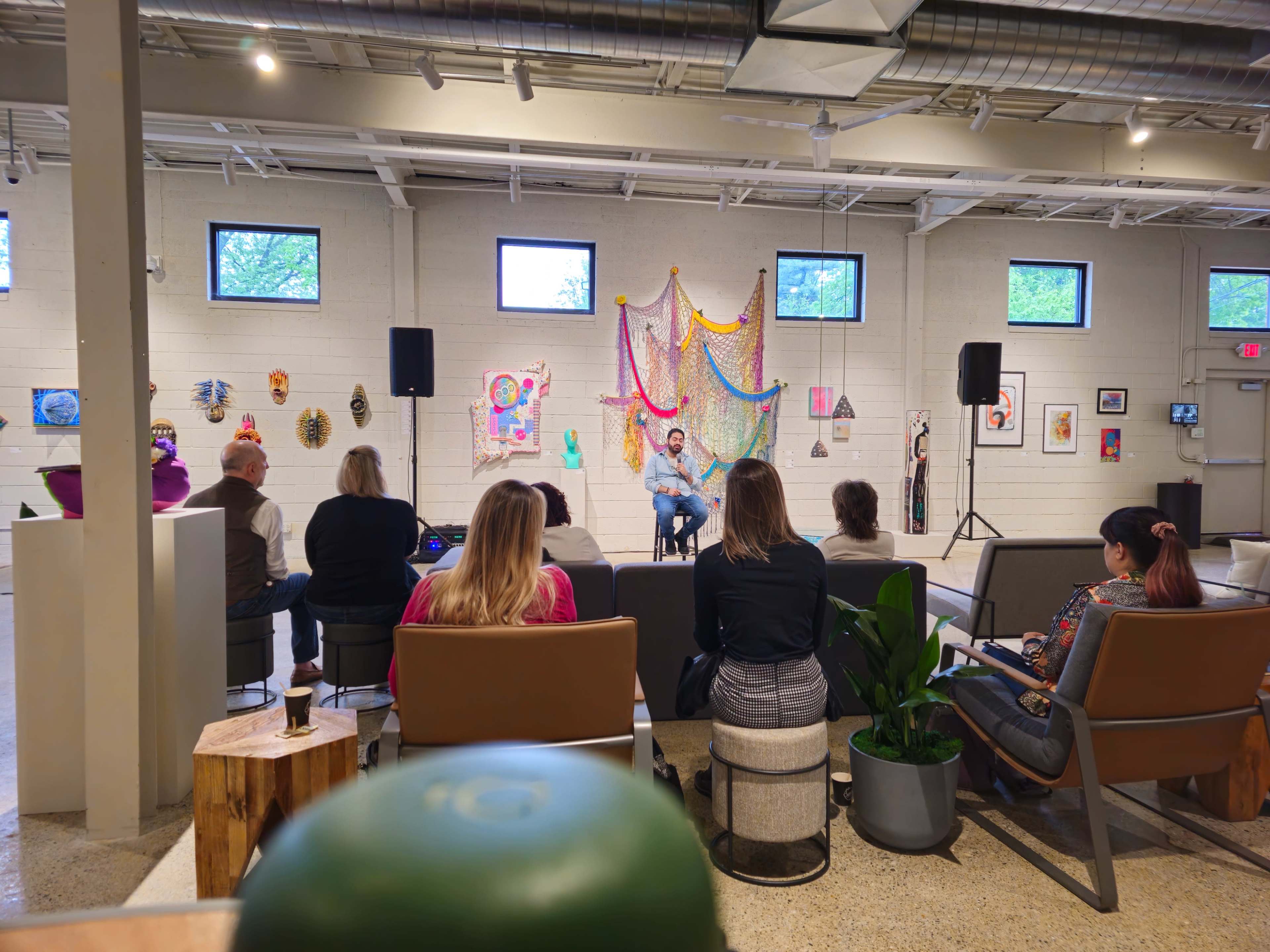 A group of people sit in a modern space with artwork displayed on the walls while a speaker presents on a stage.