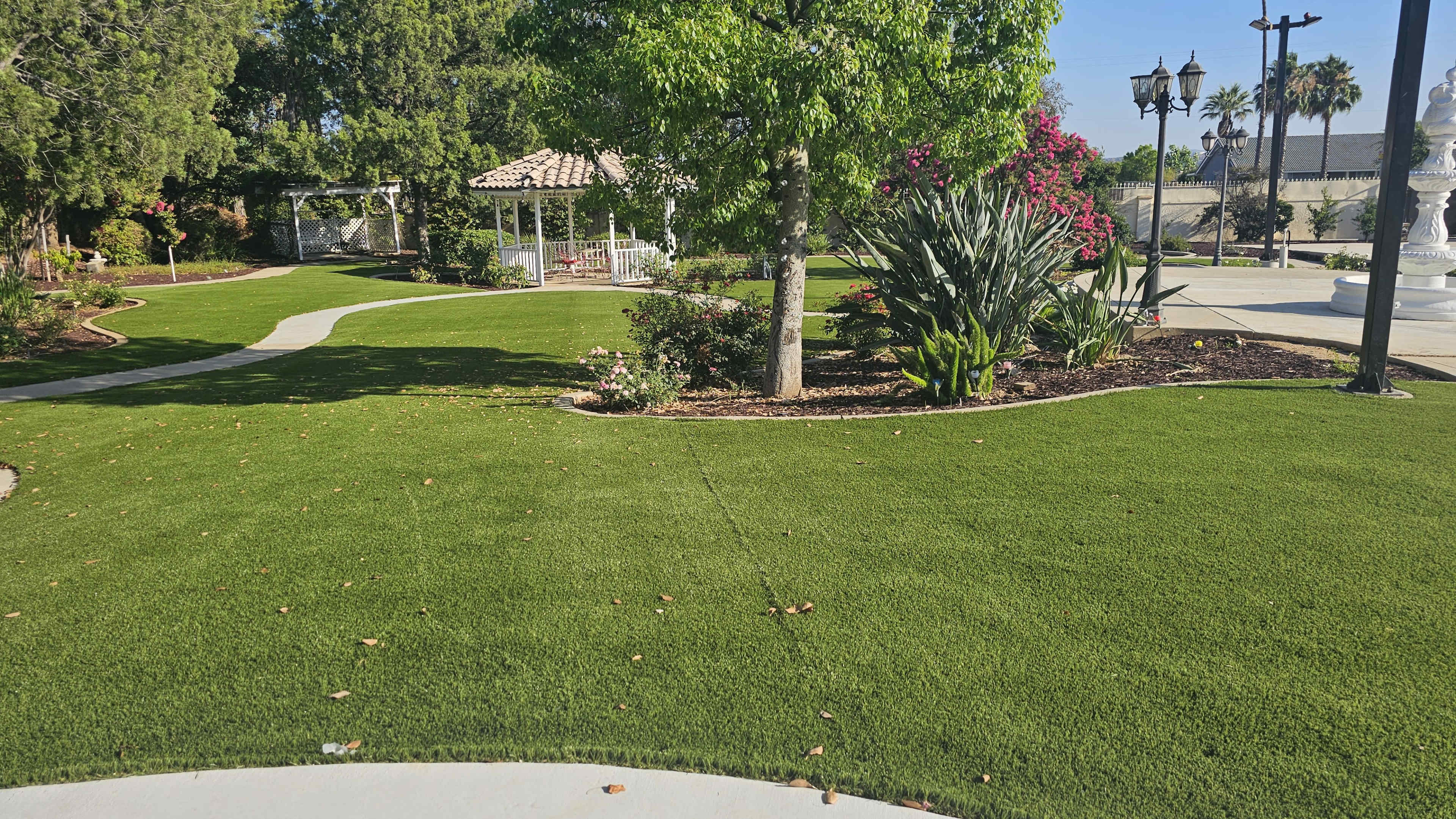 The image shows a landscaped park area with manicured green grass, a gazebo in the background, and various plants and flowers along the pathways.