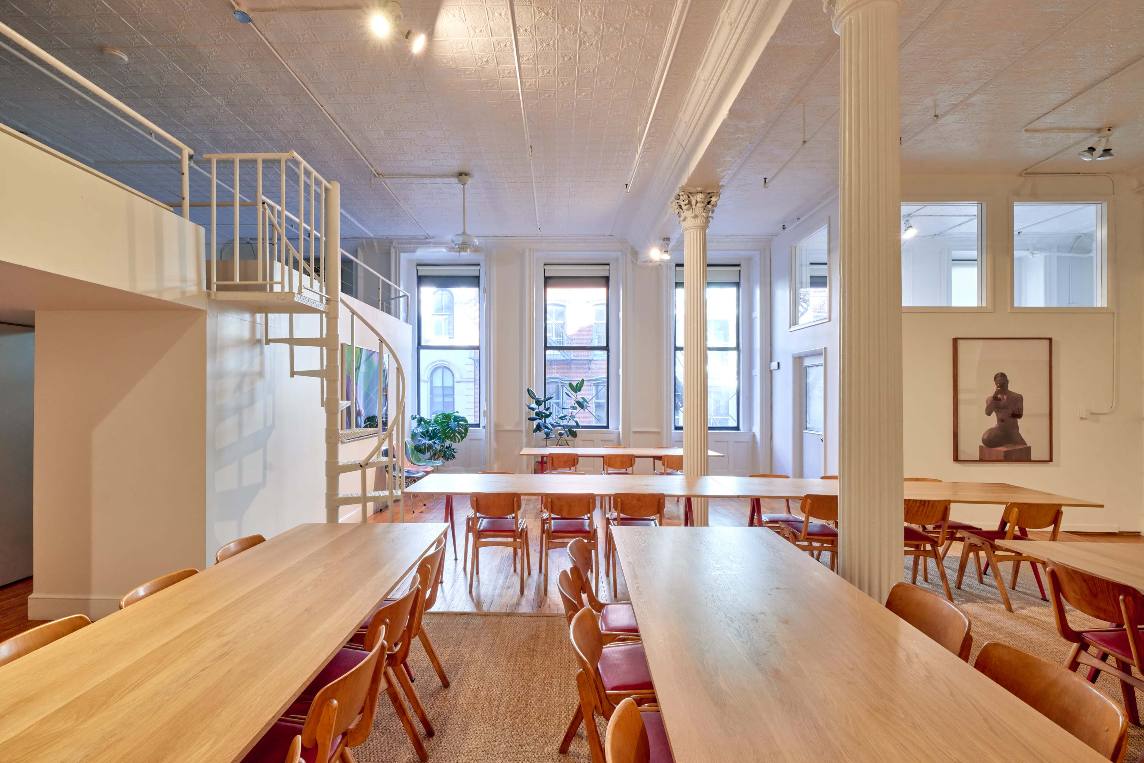 The image shows a spacious, well-lit room with multiple long wooden tables and chairs arranged for an event or meeting, featuring large windows and white pillars.