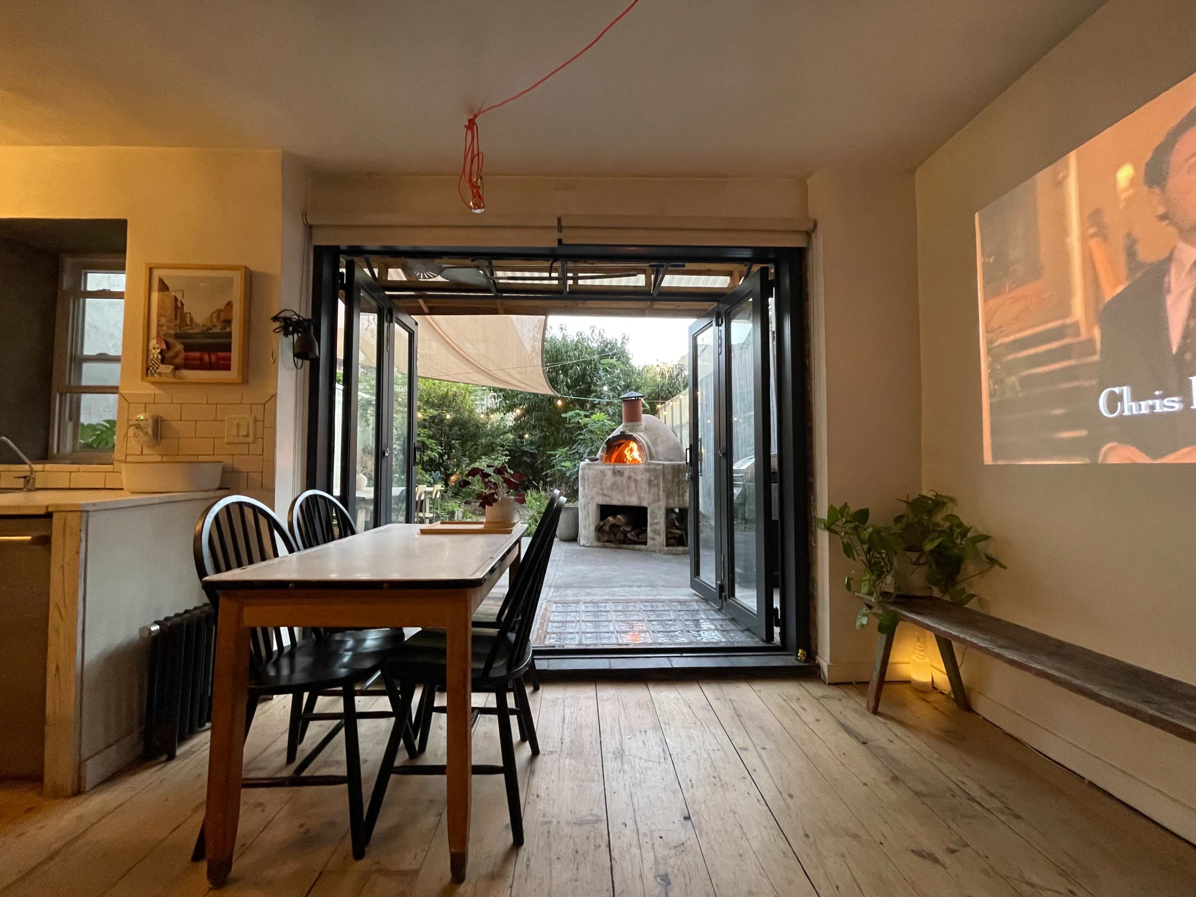 A modern dining area features a wooden table and chairs, with large open doors leading to a deck and outdoor oven.
