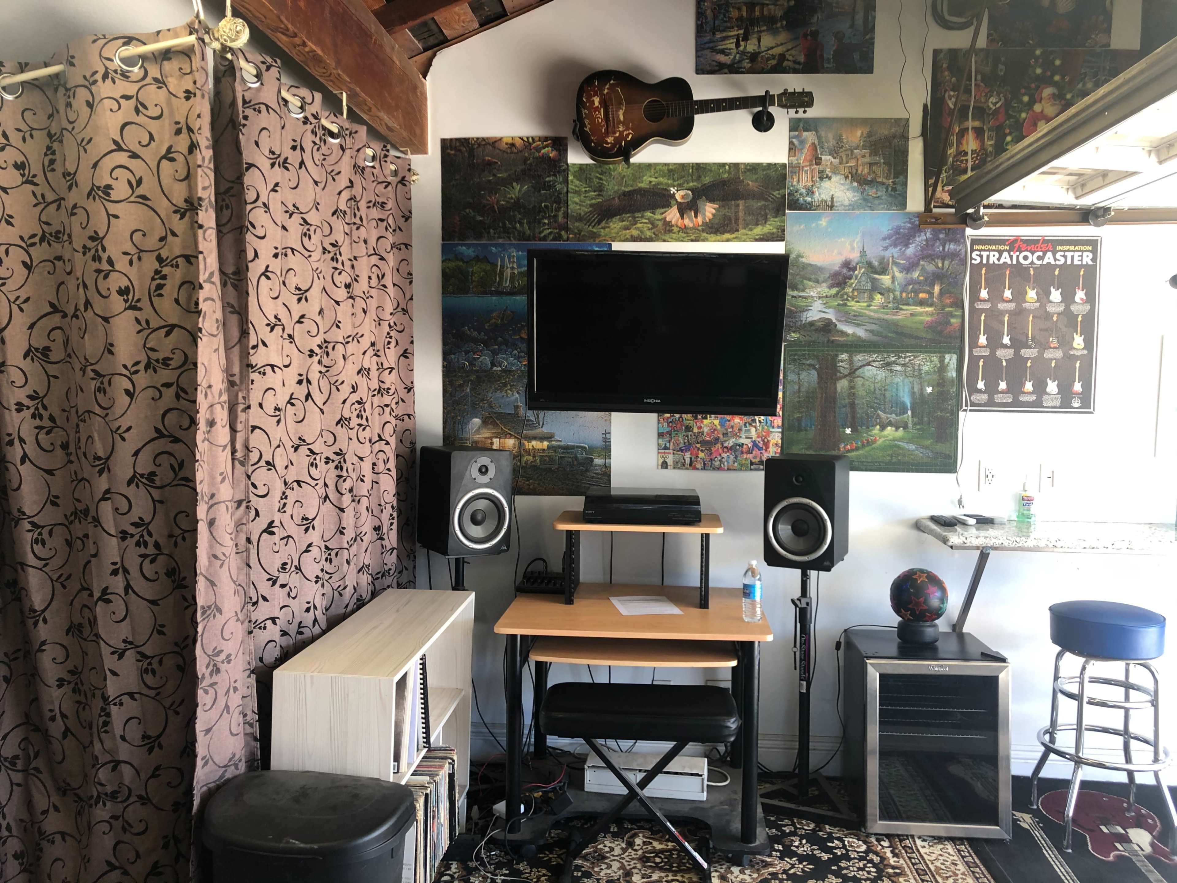 A small room featuring a television mounted on the wall, surrounded by various landscape artworks, with a music guitar hanging above, and a simple desk setup with speakers, a globe, and a water bottle.