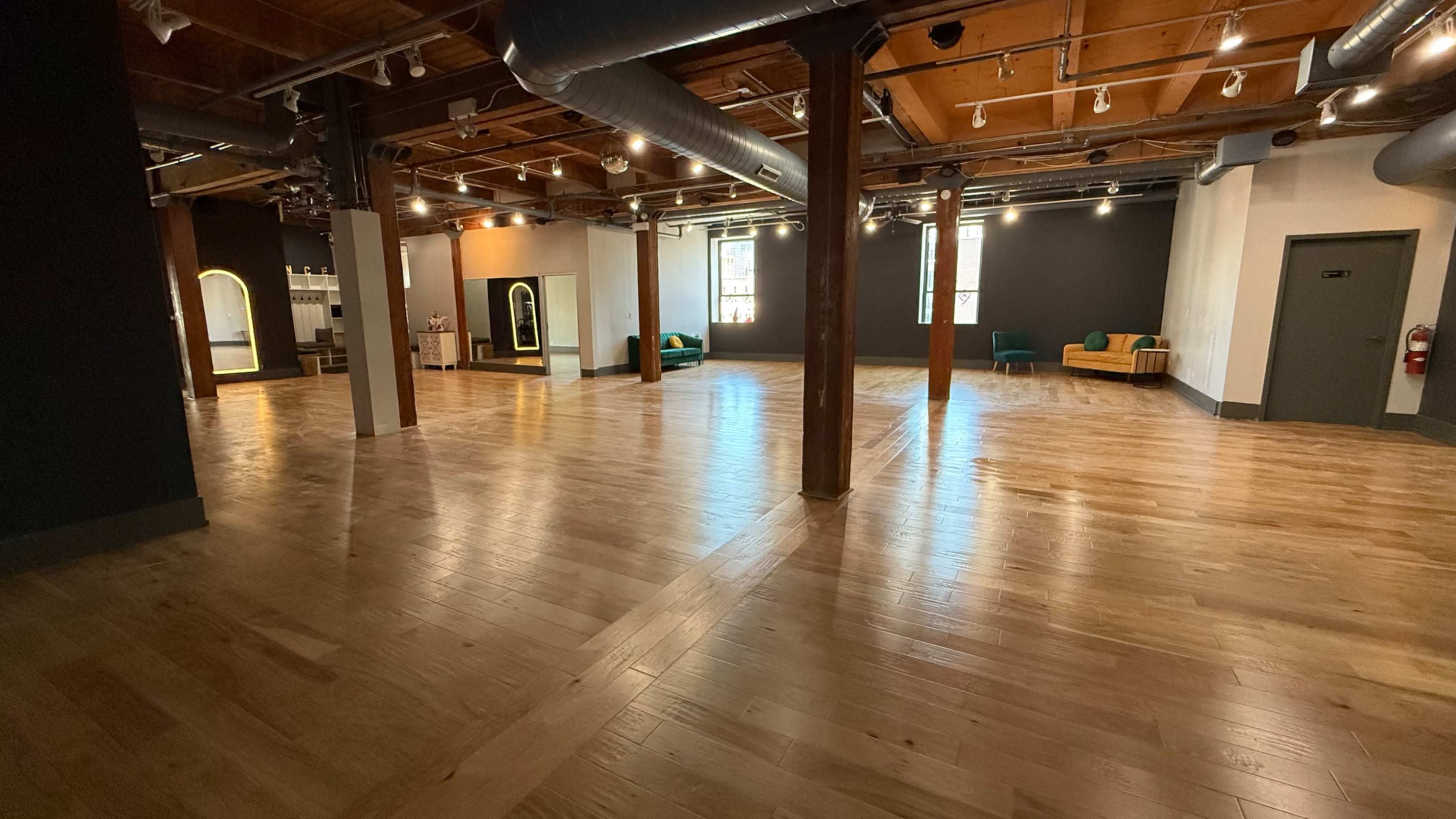 The image shows a spacious interior with polished wooden floors, large wooden pillars, and minimal furniture including two green chairs and a couch, illuminated by natural light.