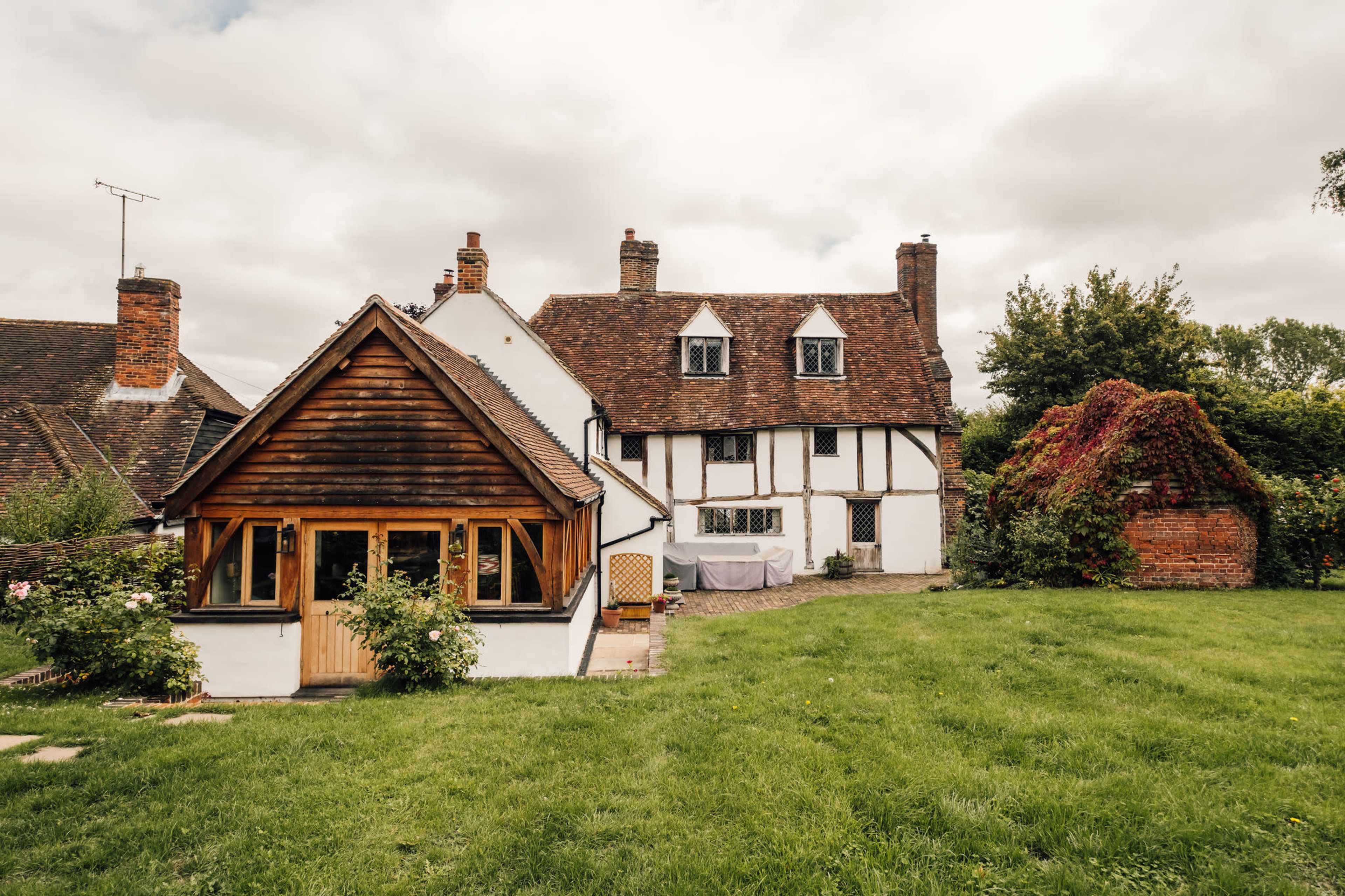 A traditional two-story house with a thatched roof and wooden accents sits next to a smaller wooden cottage in a grassy yard surrounded by trees.
